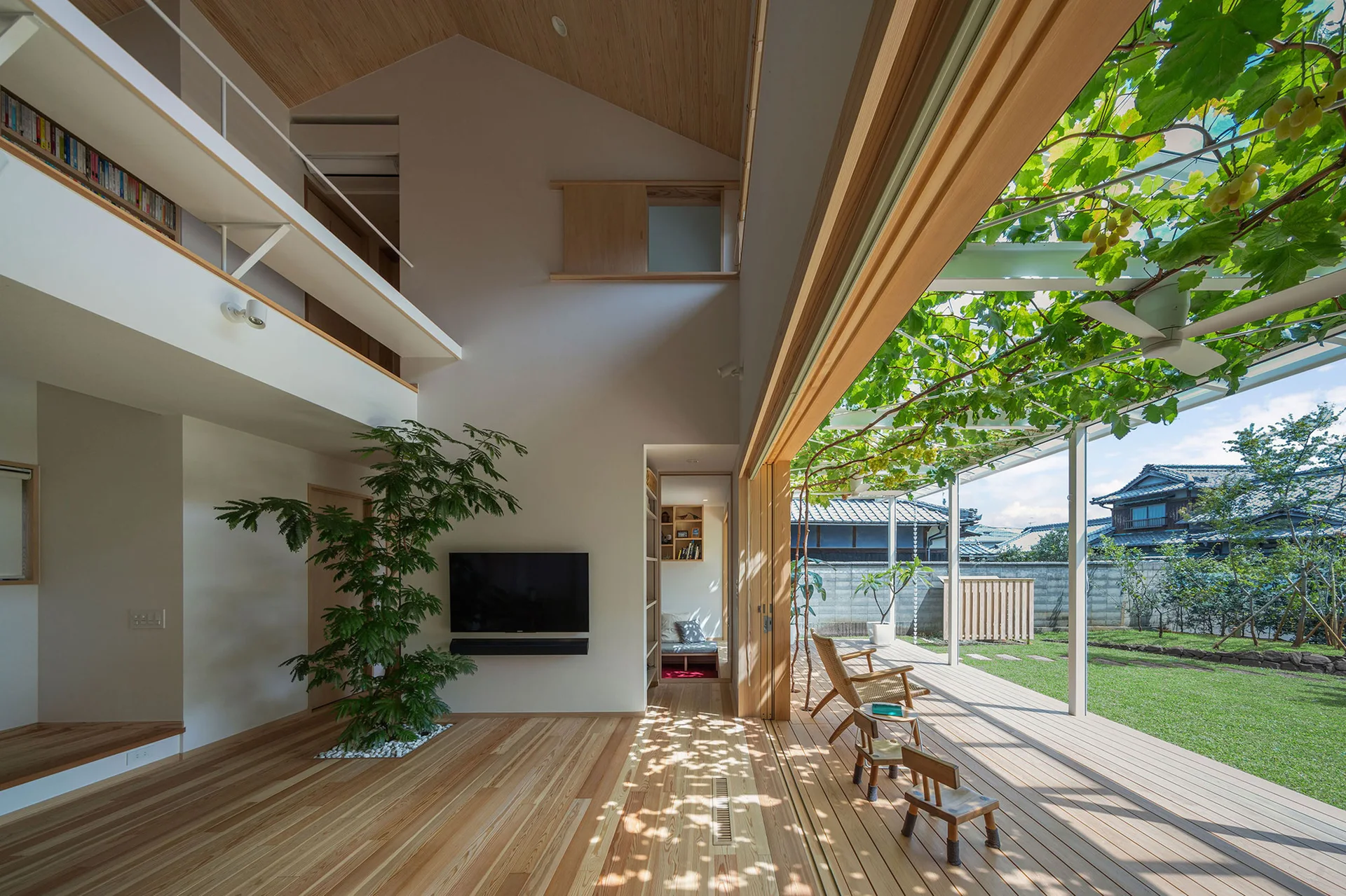 An open living room with wood floors and an indoor tree opens via sliding glass walls onto a wood deck shaded by a grapevine trellis.