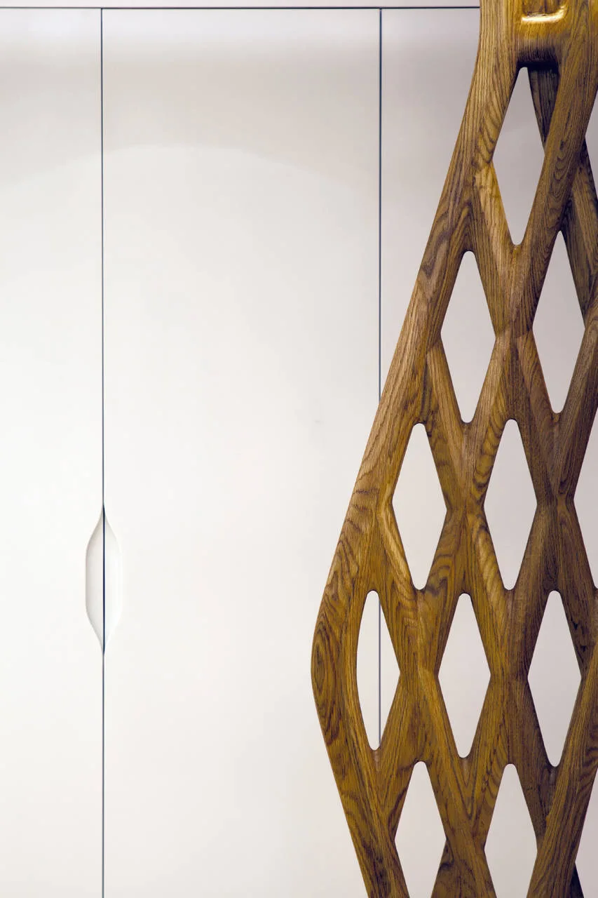 A sculptural brown wood lattice screen with diamond cutouts stands in front of flat white cabinetry with an integrated vertical handle.