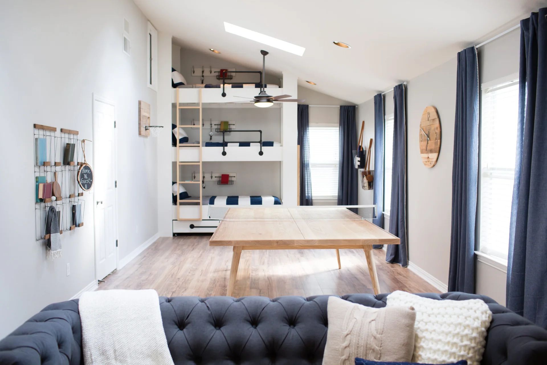 A recreation room with a built-in white triple bunk bed, a light wood ping pong table, and a grey tufted sofa in the foreground.