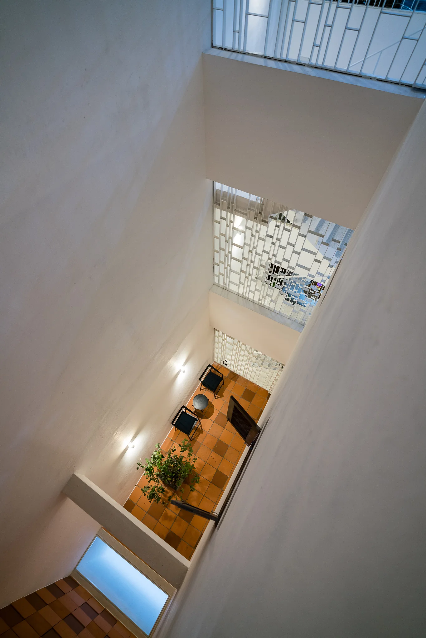 Looking down from a high angle into a narrow interior light well featuring terracotta tile flooring, two black chairs, and geometric metal screens.
