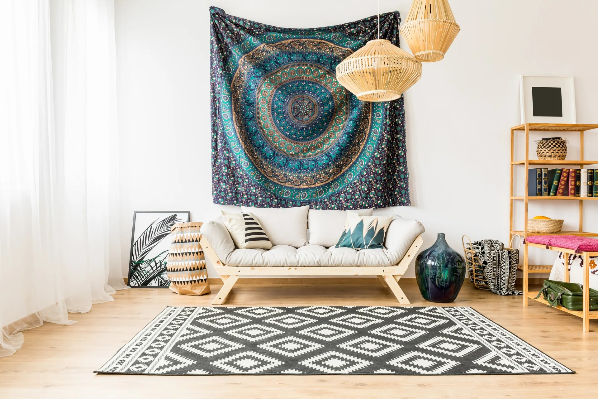 A bright living room features a wooden daybed sofa below a blue mandala tapestry, woven bamboo pendant lights, and a geometric black-and-white rug.