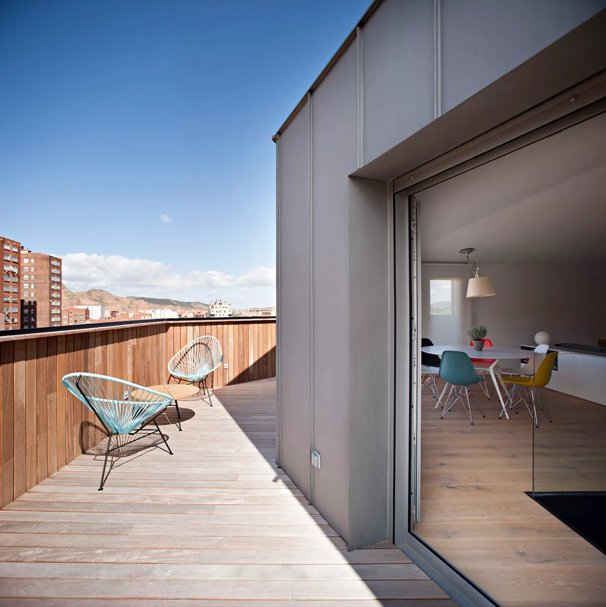 Bright blue wire chairs sit on a wood-planked terrace next to an open glass door revealing a colorful indoor dining set.