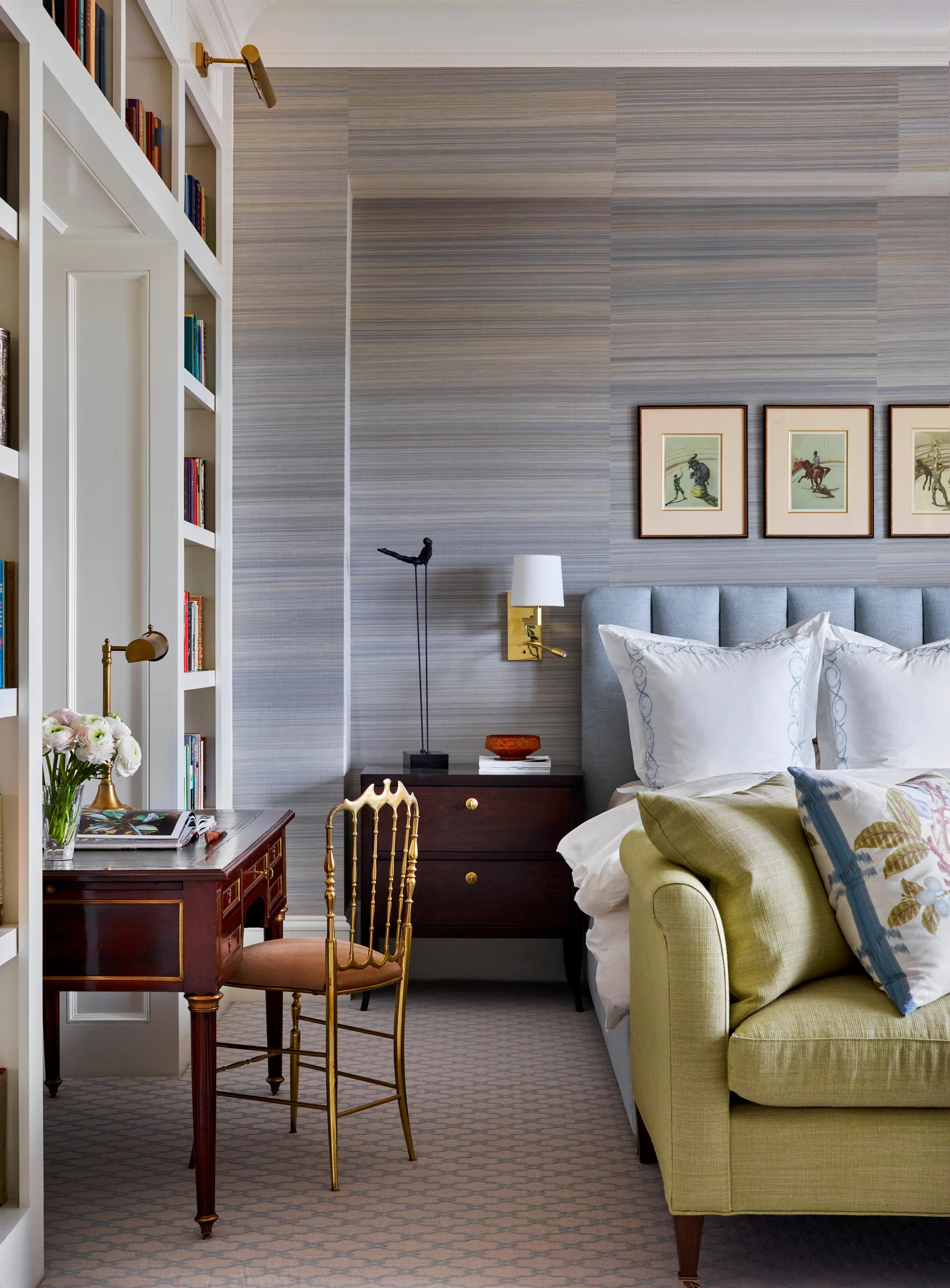 A transitional bedroom featuring a blue headboard, a dark wood writing desk with a gold chair, and textured gray grasscloth wallpaper.