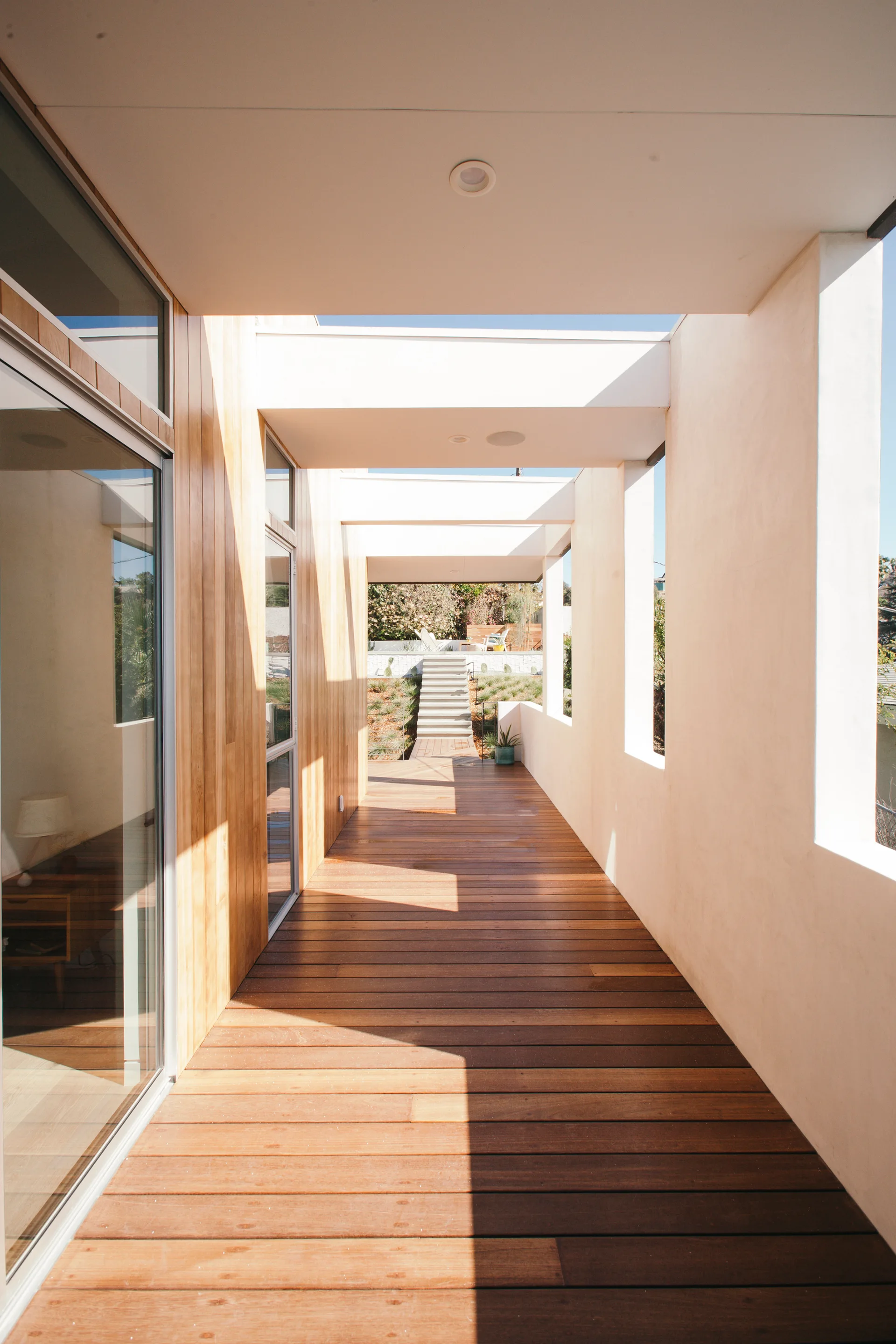 A long outdoor walkway features warm wood plank decking, a wood-clad wall with sliding glass doors, and white stucco walls with overhead beams.