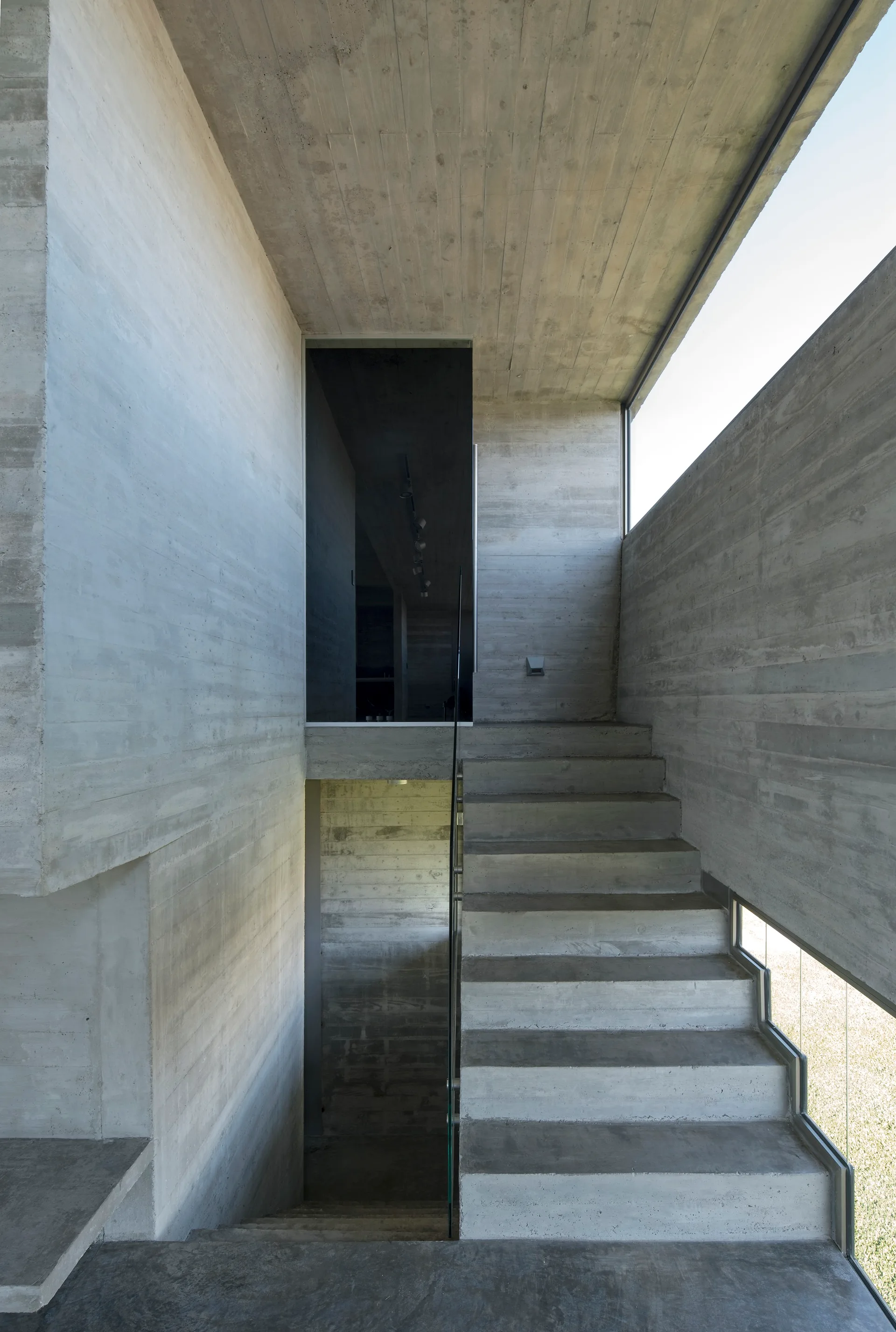 A multi-level concrete staircase features board-formed concrete walls, a transparent glass railing, and narrow horizontal windows.