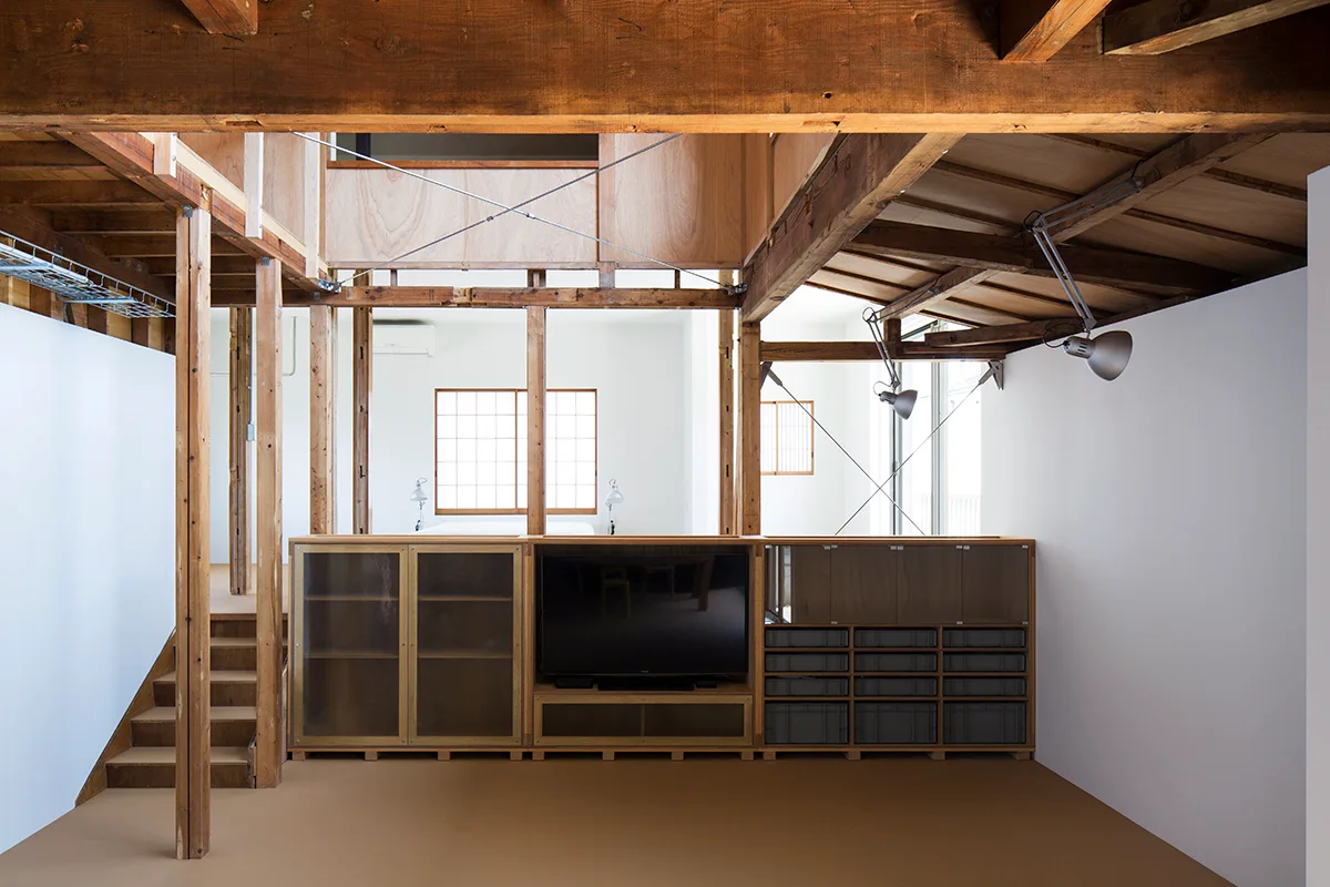 A modern living space features exposed wooden ceiling beams, white walls, and a low custom wood cabinet with a television and frosted glass doors.