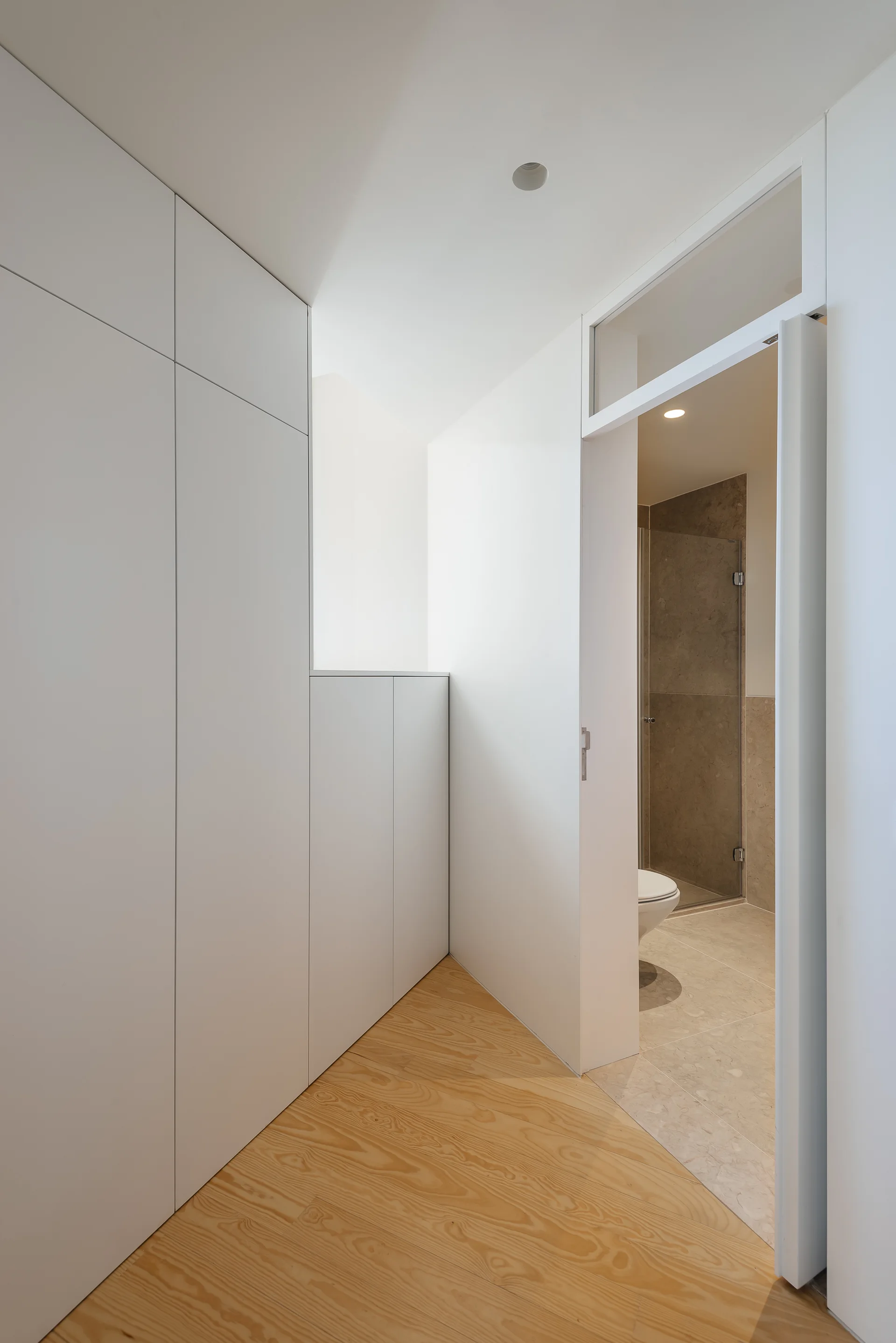 A minimalist white hallway with floor-to-ceiling built-in cabinets and light wood floors leads to a tiled bathroom with a glass transom.