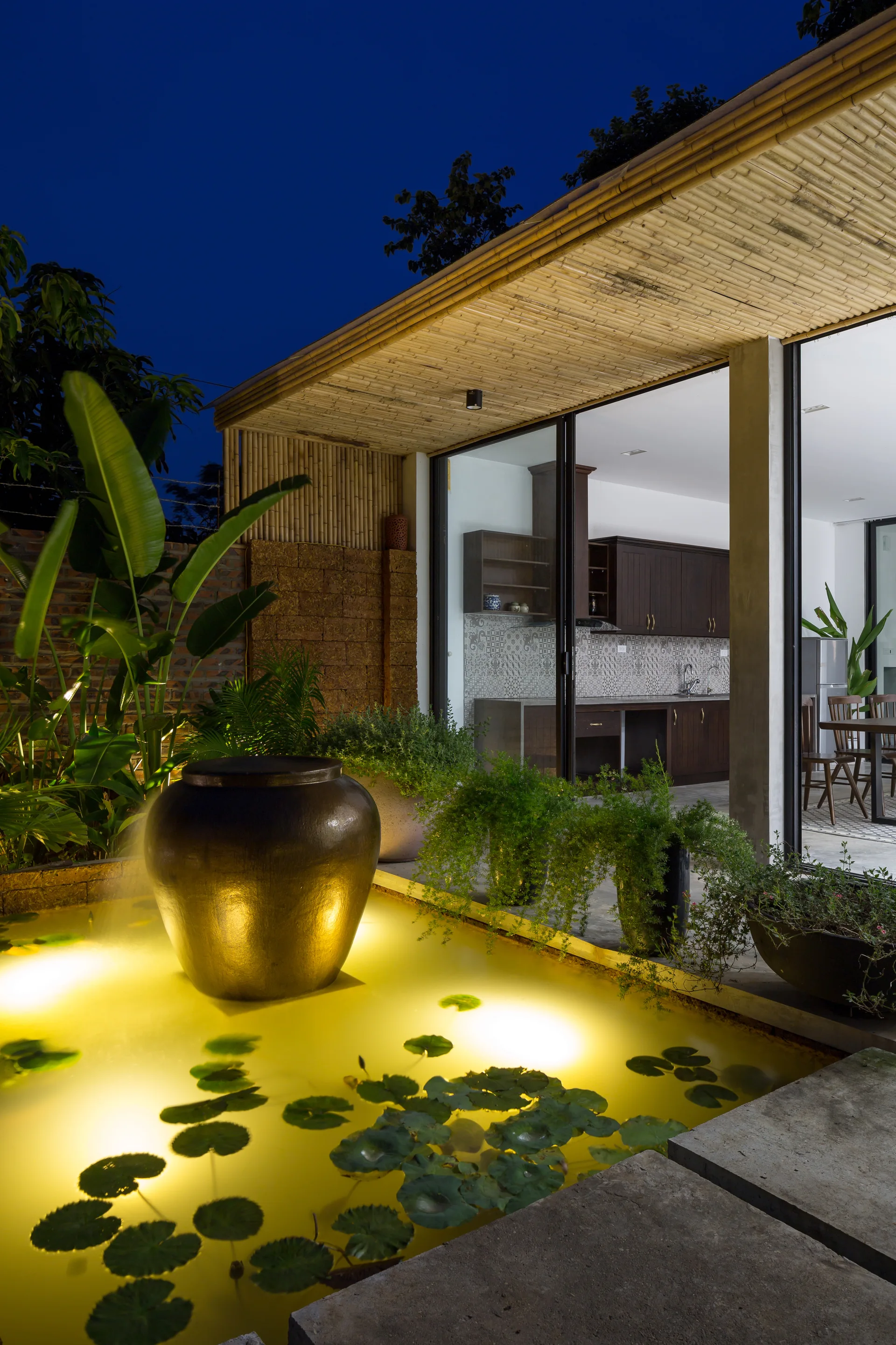 A night view of an outdoor patio featuring an illuminated pond with lily pads, a large urn fountain, and a bamboo roof overhang.