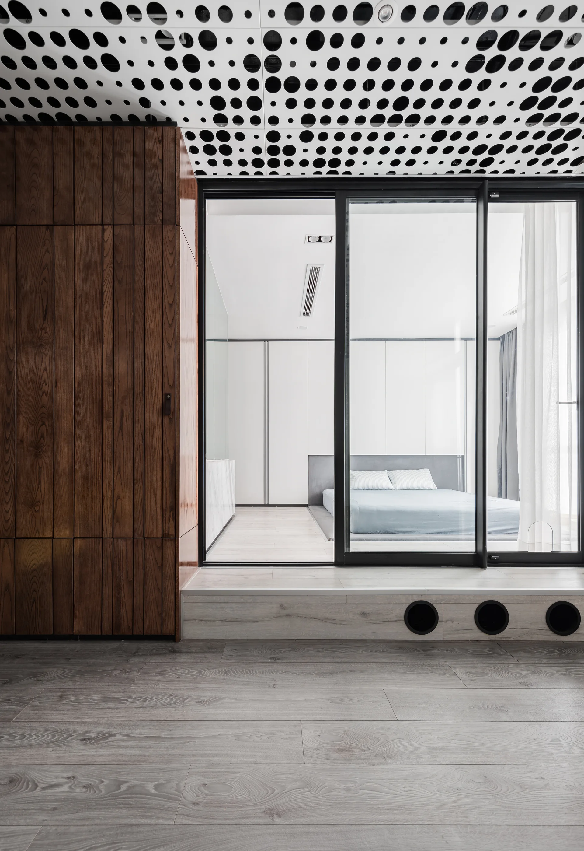 A modern bedroom with a low profile bed on a raised platform, seen through a black-framed sliding glass door next to a wood cabinet.
