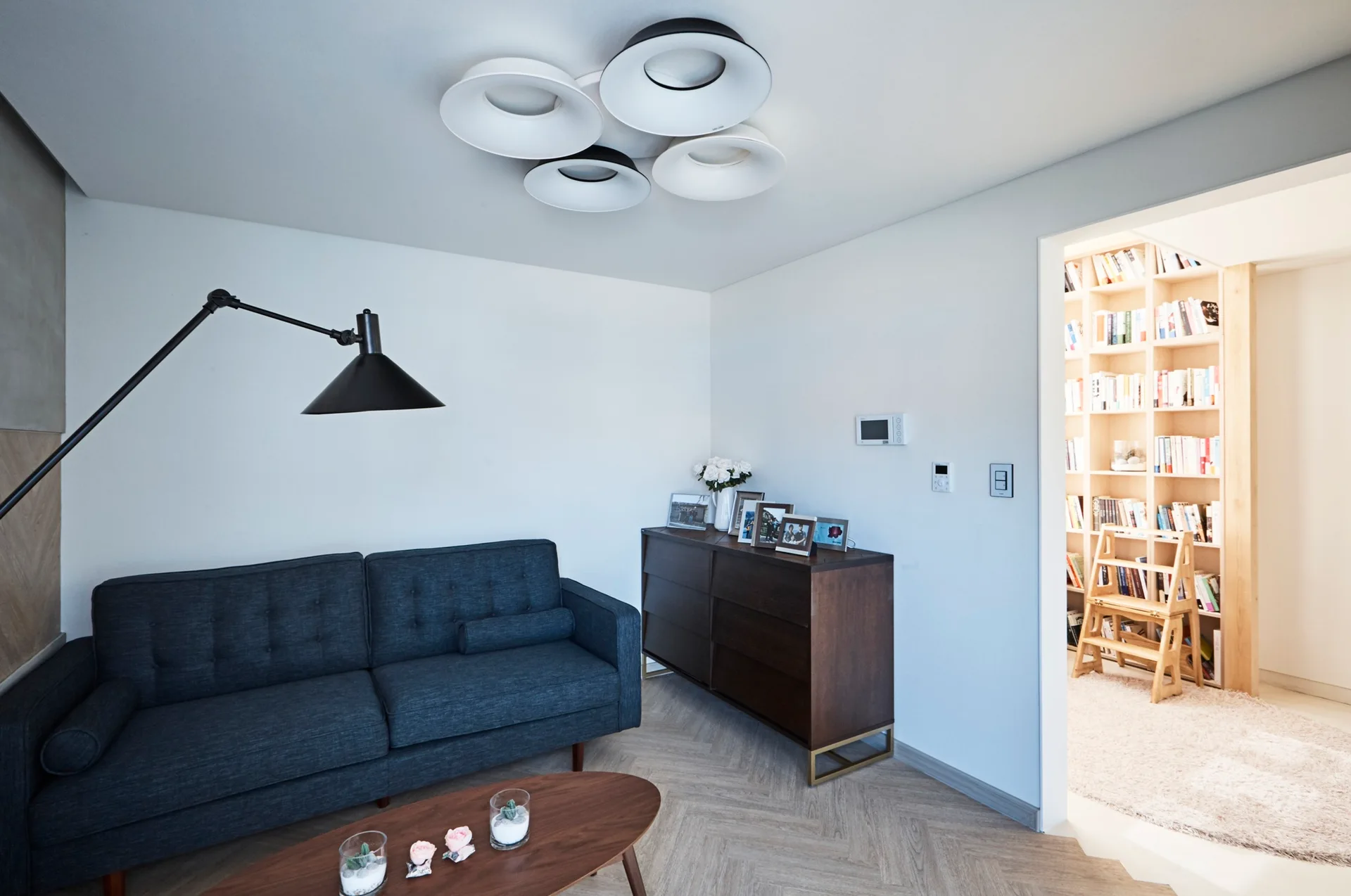A living room features a charcoal gray sofa, herringbone wood floors, an articulating black lamp, and an open doorway leading to a home library.