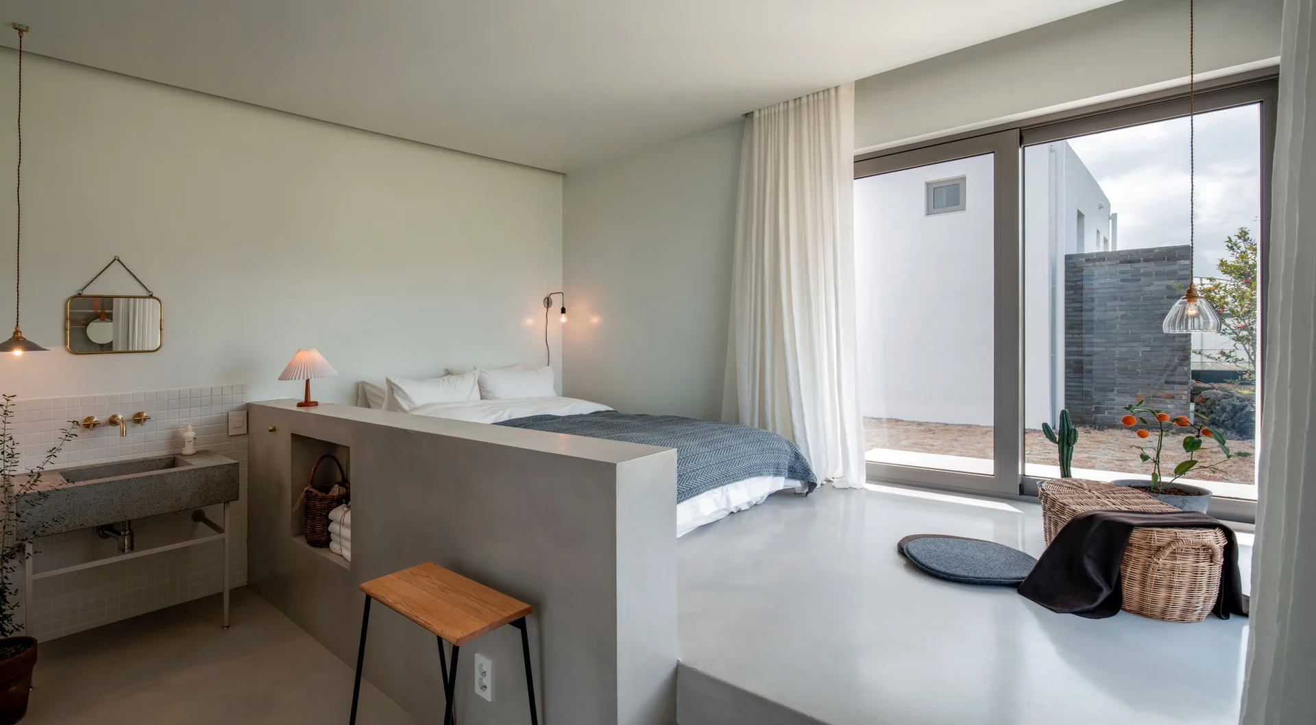 A minimalist bedroom features a low concrete partition wall dividing the bed from a stone vanity sink, next to large glass doors.