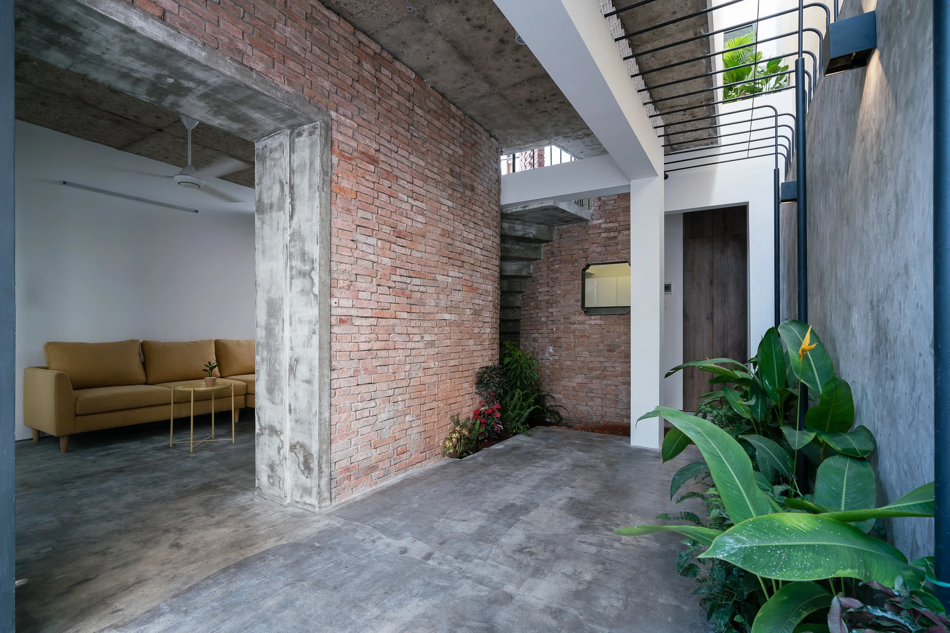 An industrial interior space features exposed red brick walls, raw concrete surfaces, an indoor plant bed, and a living area with a mustard yellow sofa.