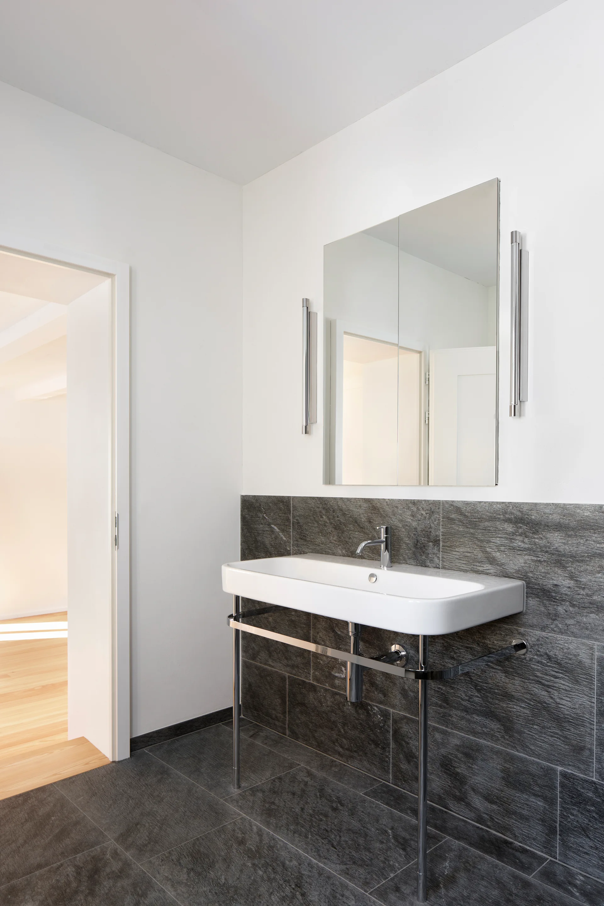 A white washbasin with polished chrome legs stands against dark grey stone wall tiles in a minimalist bathroom with a rectangular mirror.