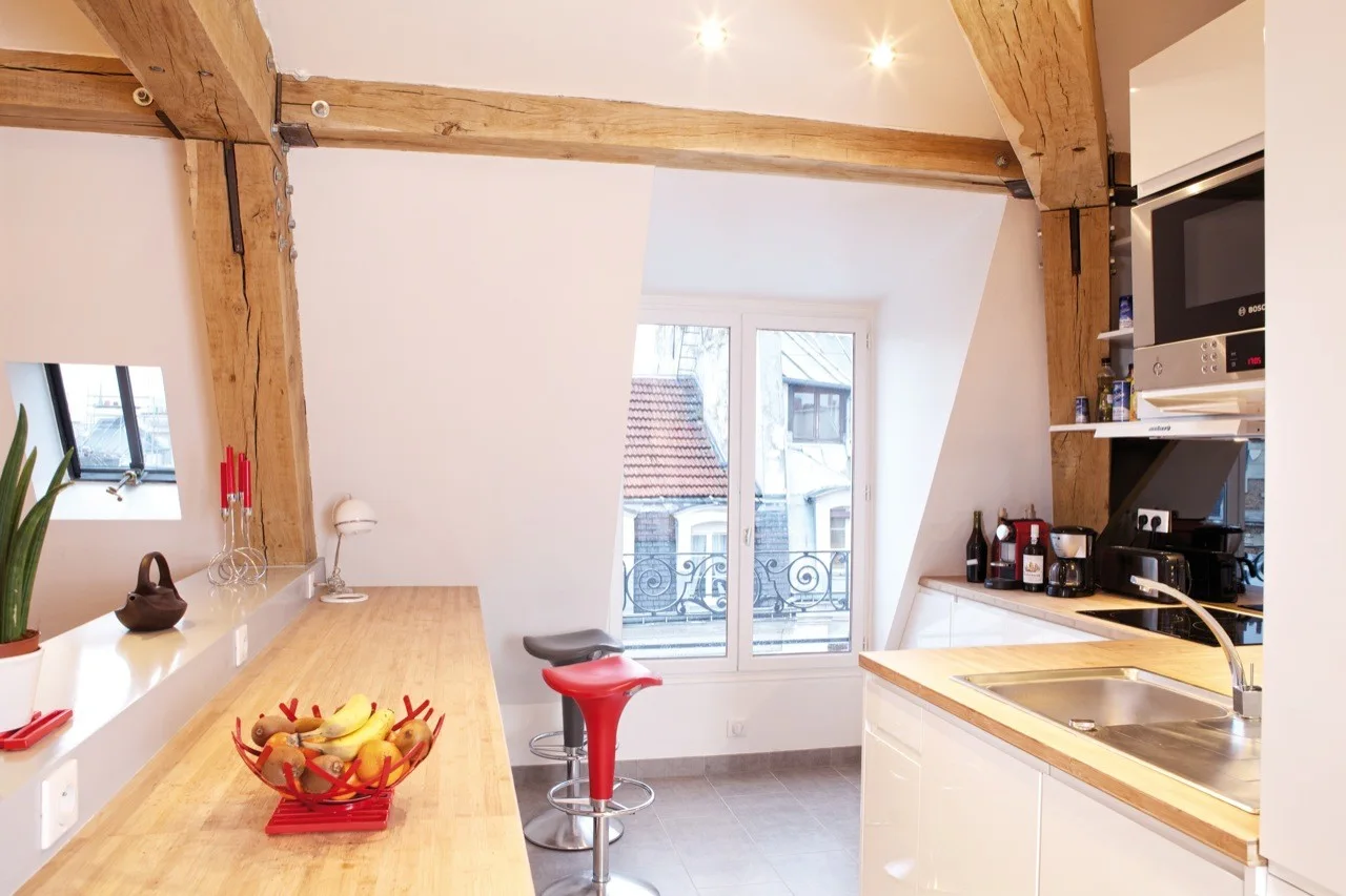 A bright attic kitchen featuring exposed wooden ceiling beams, white cabinetry, a wood-topped breakfast bar with red and grey stools, and a window.