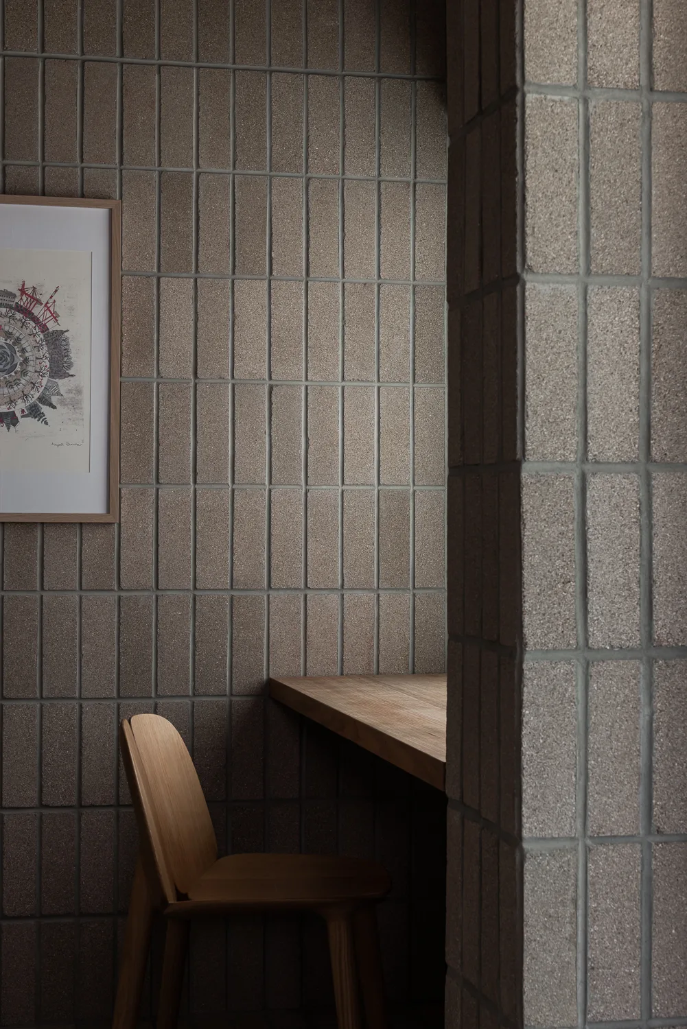 A minimalist wooden desk and chair sit nestled against a wall of vertically stacked taupe tiles next to a structural pillar.
