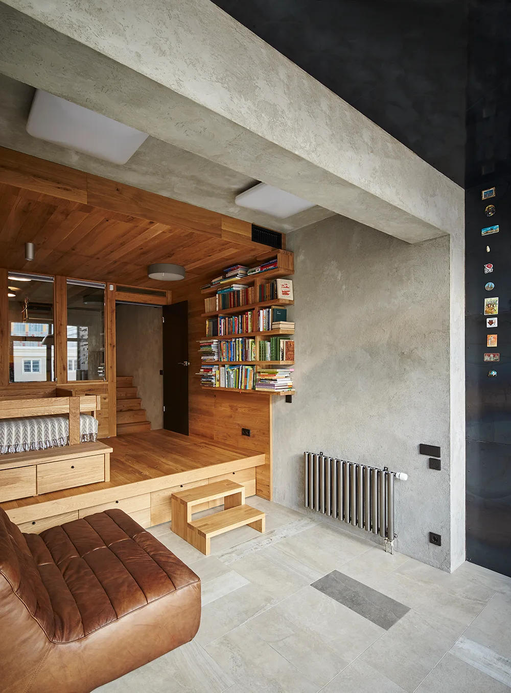 A modern residential space features a raised light wood platform with integrated drawers, concrete walls, and a brown leather lounge chair.