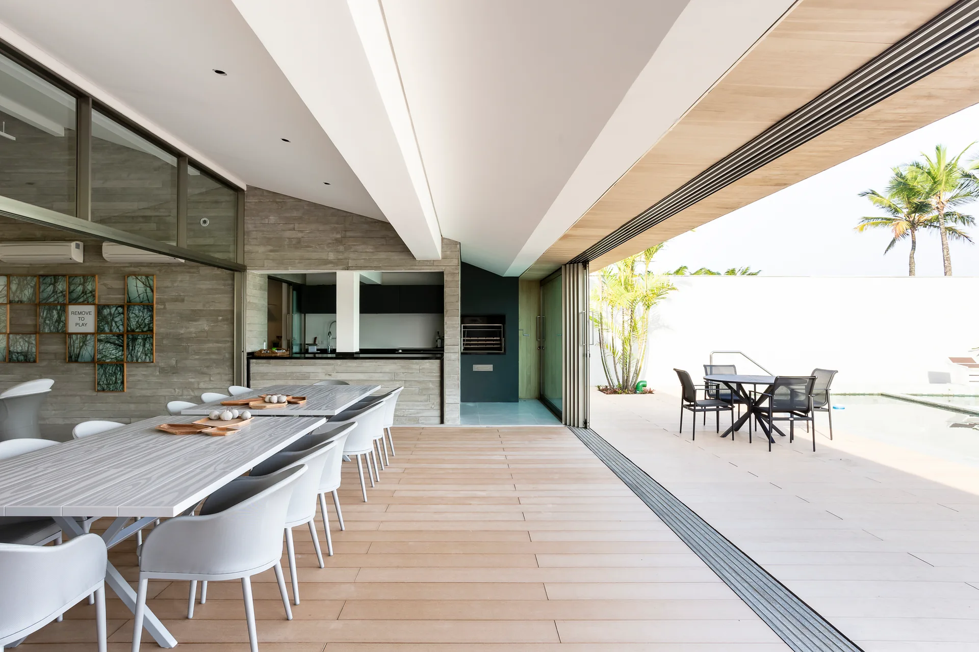 A covered patio dining area with long white tables and chairs features a wood-look plank floor that extends toward an outdoor swimming pool.