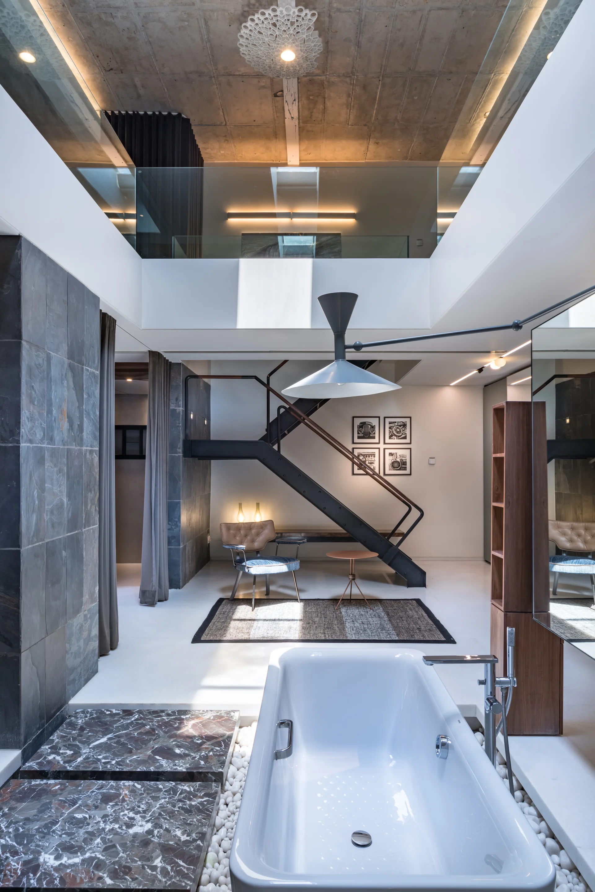 A contemporary open-concept bathroom features a white soaking tub bordered by pebbles next to a black metal staircase and slate tile wall.