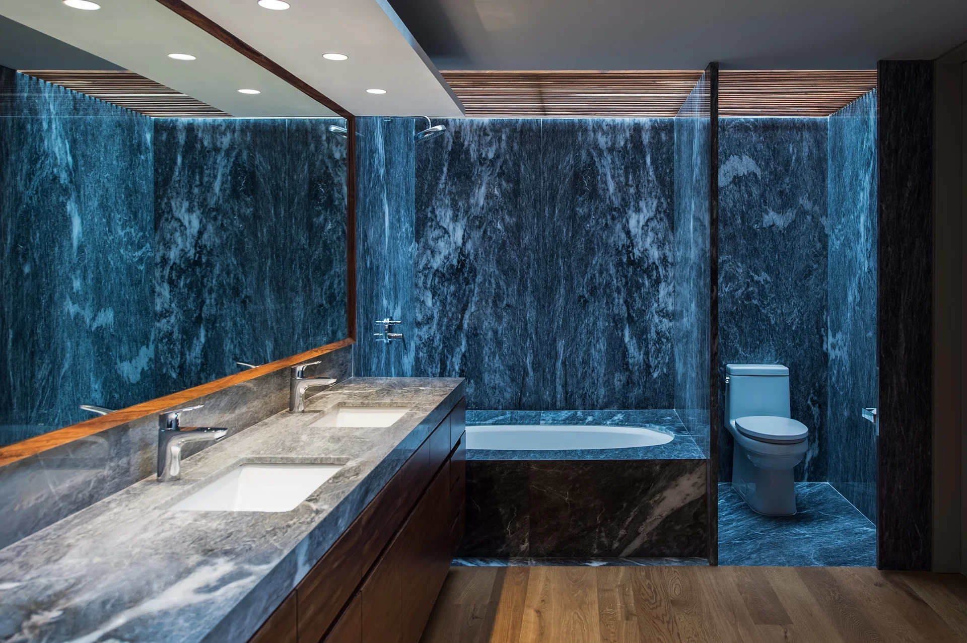 A modern bathroom with dark grey veined marble walls, a wood double vanity, integrated ceiling lights, and wooden floor panels.