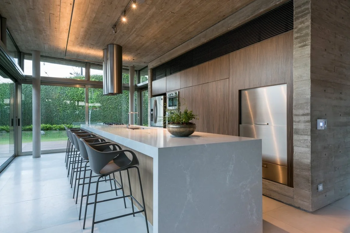 A modern kitchen features a long white waterfall island with grey bar stools, set against a wood cabinet wall and an exposed concrete ceiling.