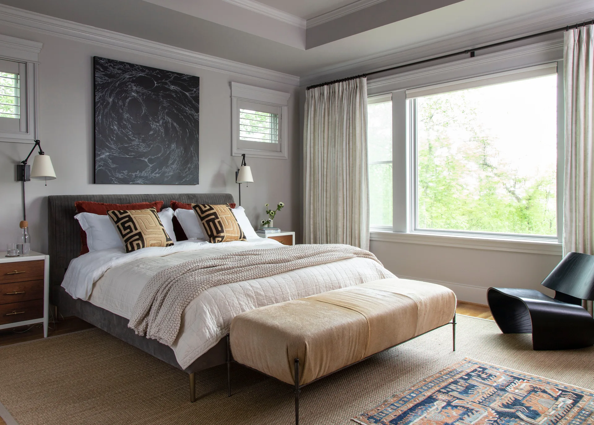 A contemporary bedroom features an upholstered gray bed, textured pillows, a leather bench, and a large black abstract painting on the wall.