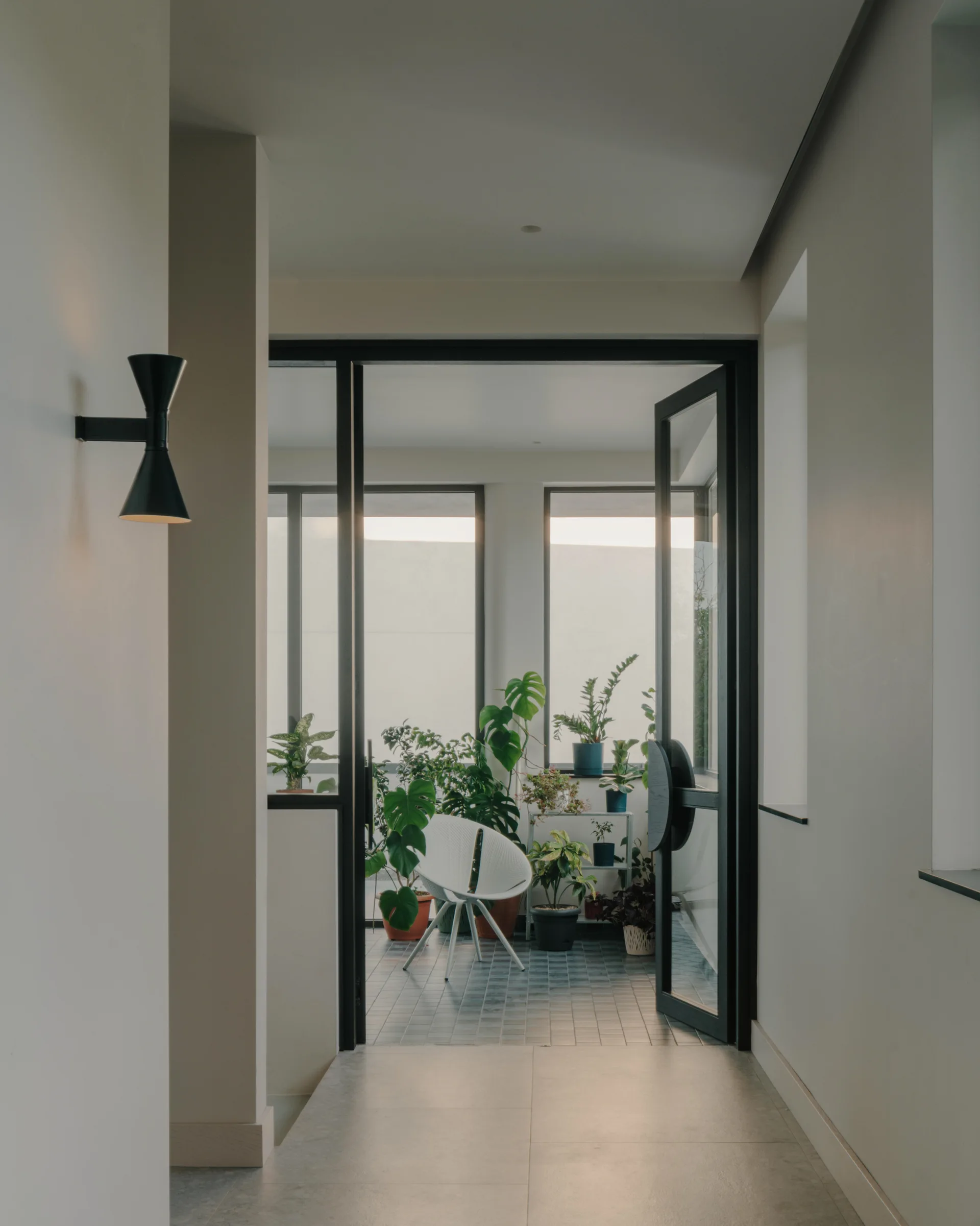 A view from a grey-walled hallway leading through black metal glass doors into a bright terrace filled with potted plants and a white chair.