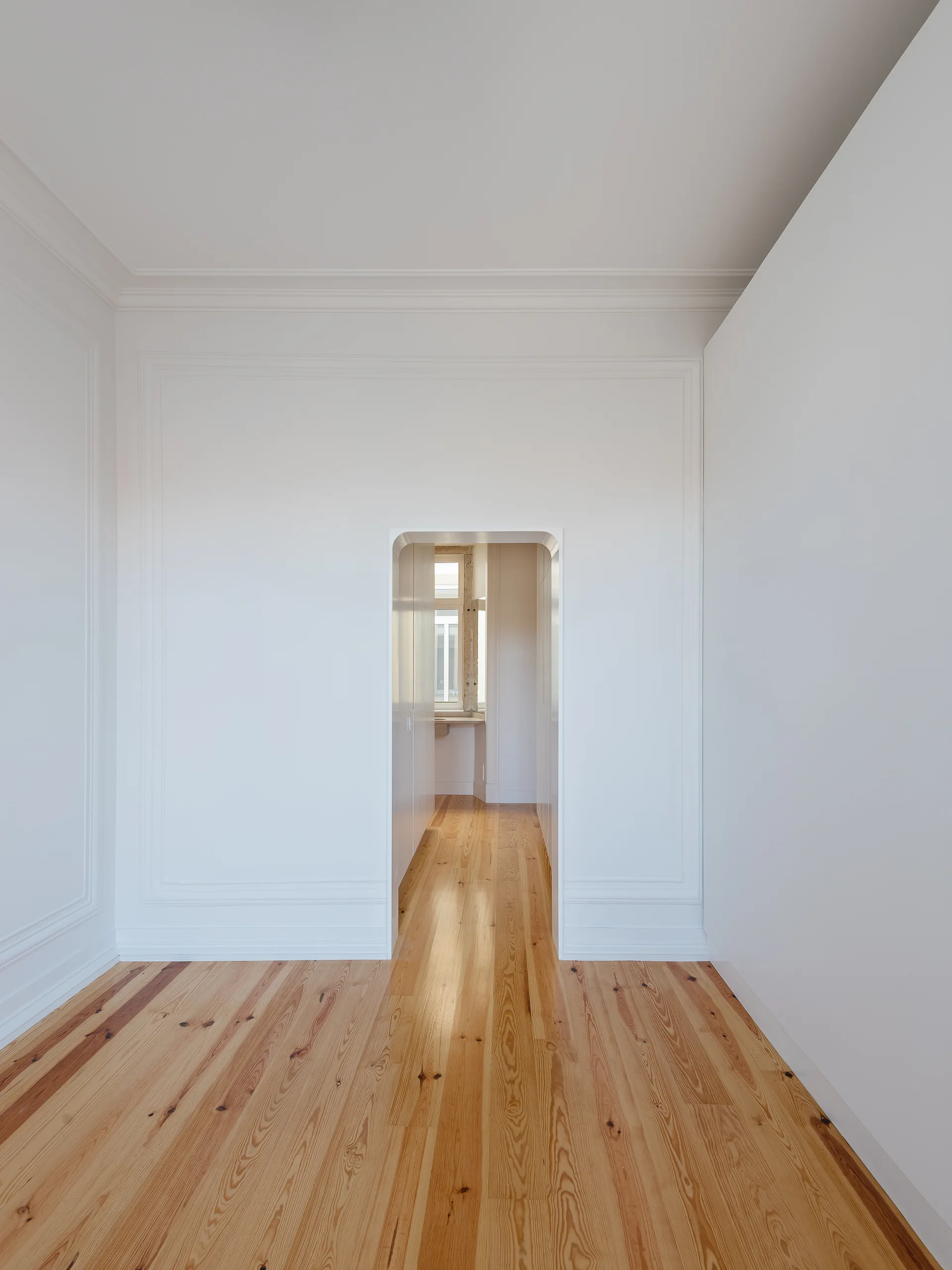 A minimalist white room with wall molding, light wood floors, and a central doorway with rounded corners leading to a hallway.