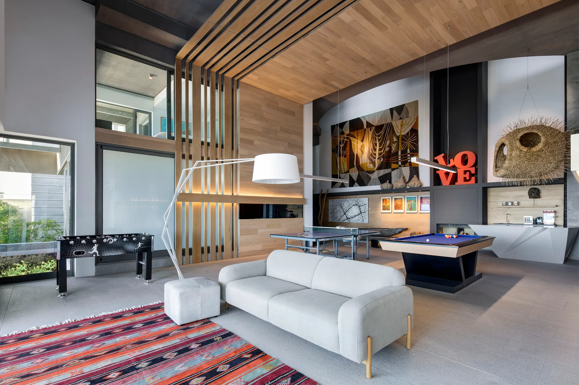 A contemporary open-concept game room featuring a light grey sofa, a blue pool table, a foosball table, and a wood-slat ceiling and wall canopy.