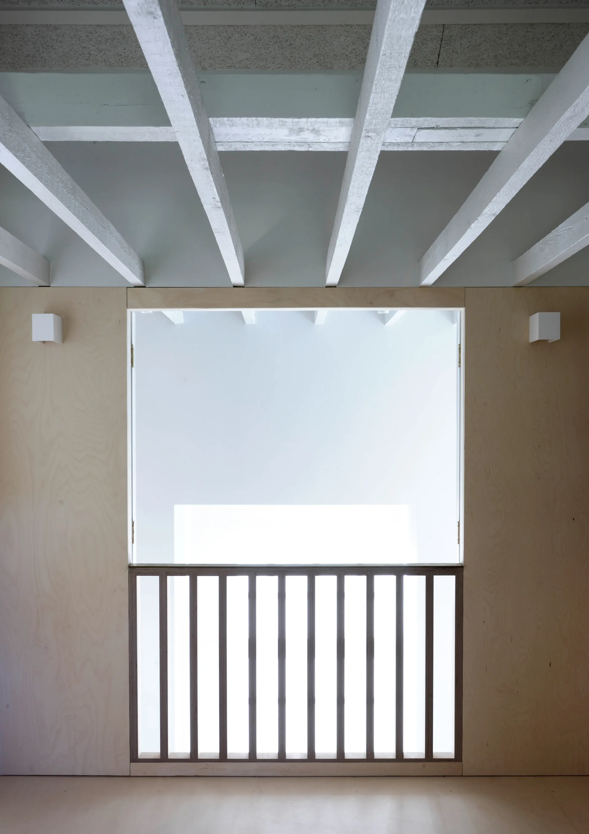 A minimalist mezzanine landing features light plywood wall paneling, a dark wood slatted railing, and an exposed white beam ceiling.