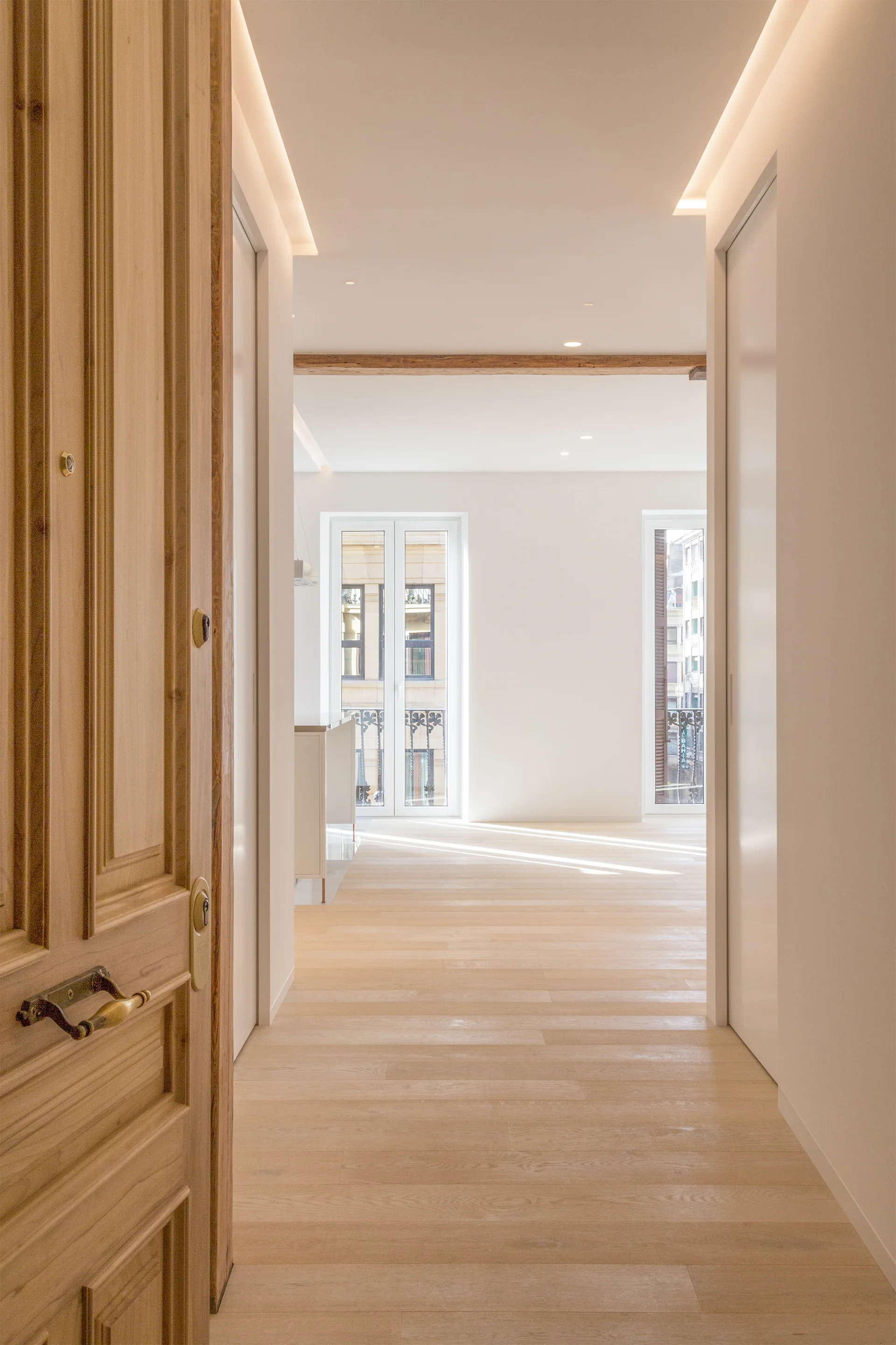 A light wood door opens into a bright minimalist hallway featuring wood plank floors, clean white walls, and built-in ceiling cove lighting.