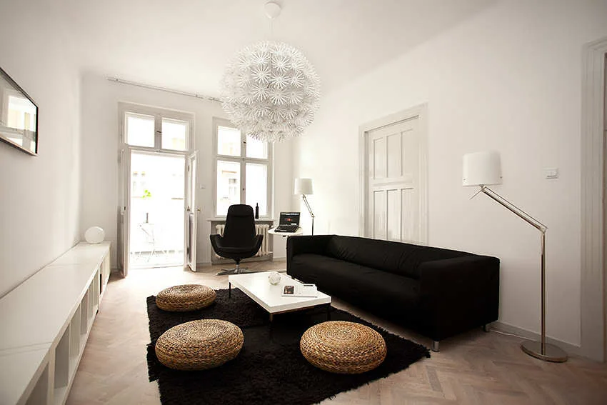 A minimalist living room featuring a black sofa, woven wicker poufs, and a large spiky spherical pendant light over herringbone parquet flooring.