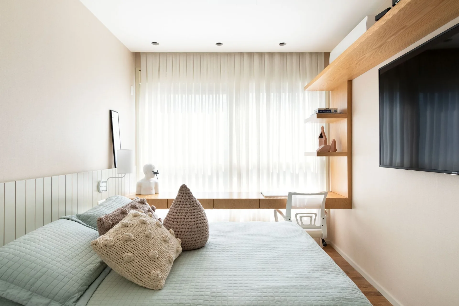 A light blue bed with textured pillows rests beside a built-in wooden floating desk and shelves under a window with sheer white curtains.