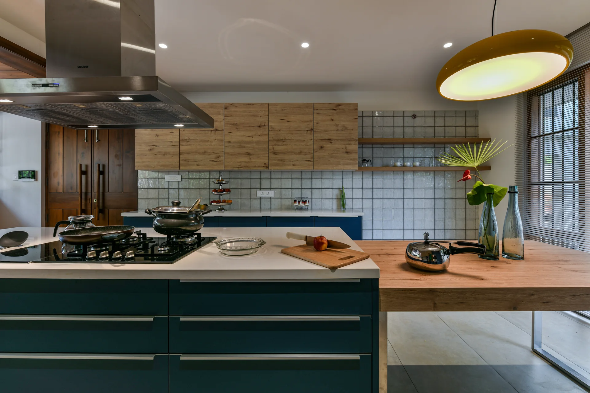 A modern kitchen featuring a navy blue island with a built-in cooktop, a wooden table extension, and wood-grained upper cabinets against a grid-tiled wall.