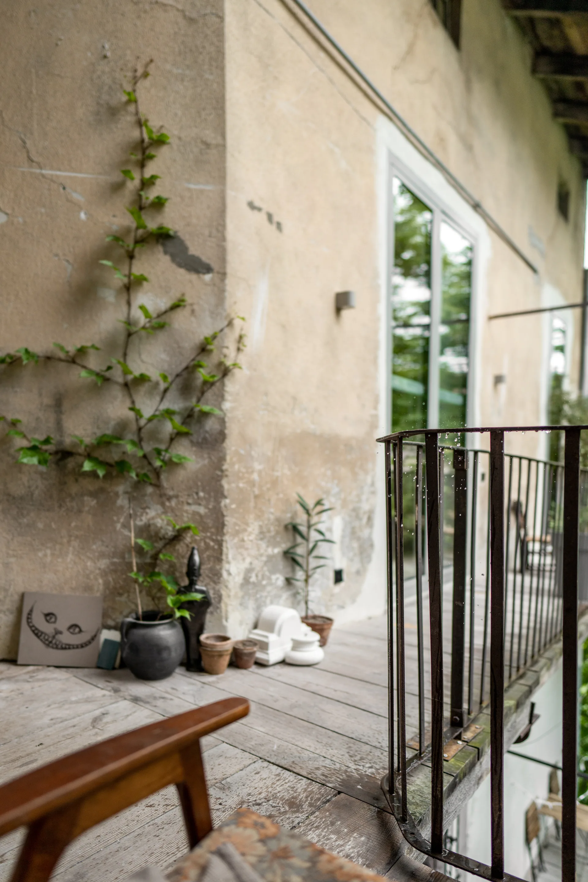 An outdoor wooden balcony with a weathered stucco wall features a climbing green vine, small potted plants, and a dark metal railing.
