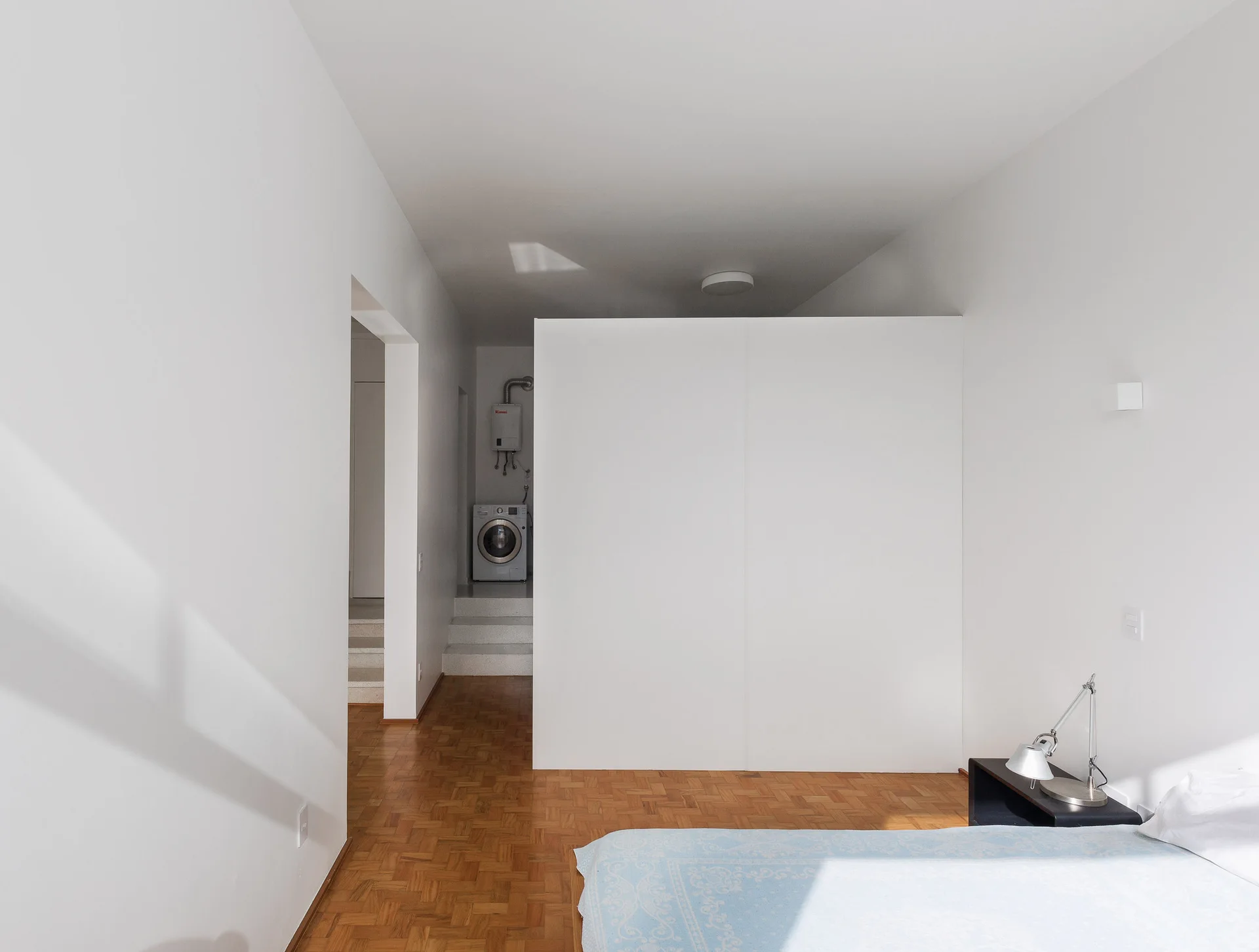 A minimalist bedroom featuring warm wood parquet flooring, a large white partition wall, and a view of steps leading to a laundry area.