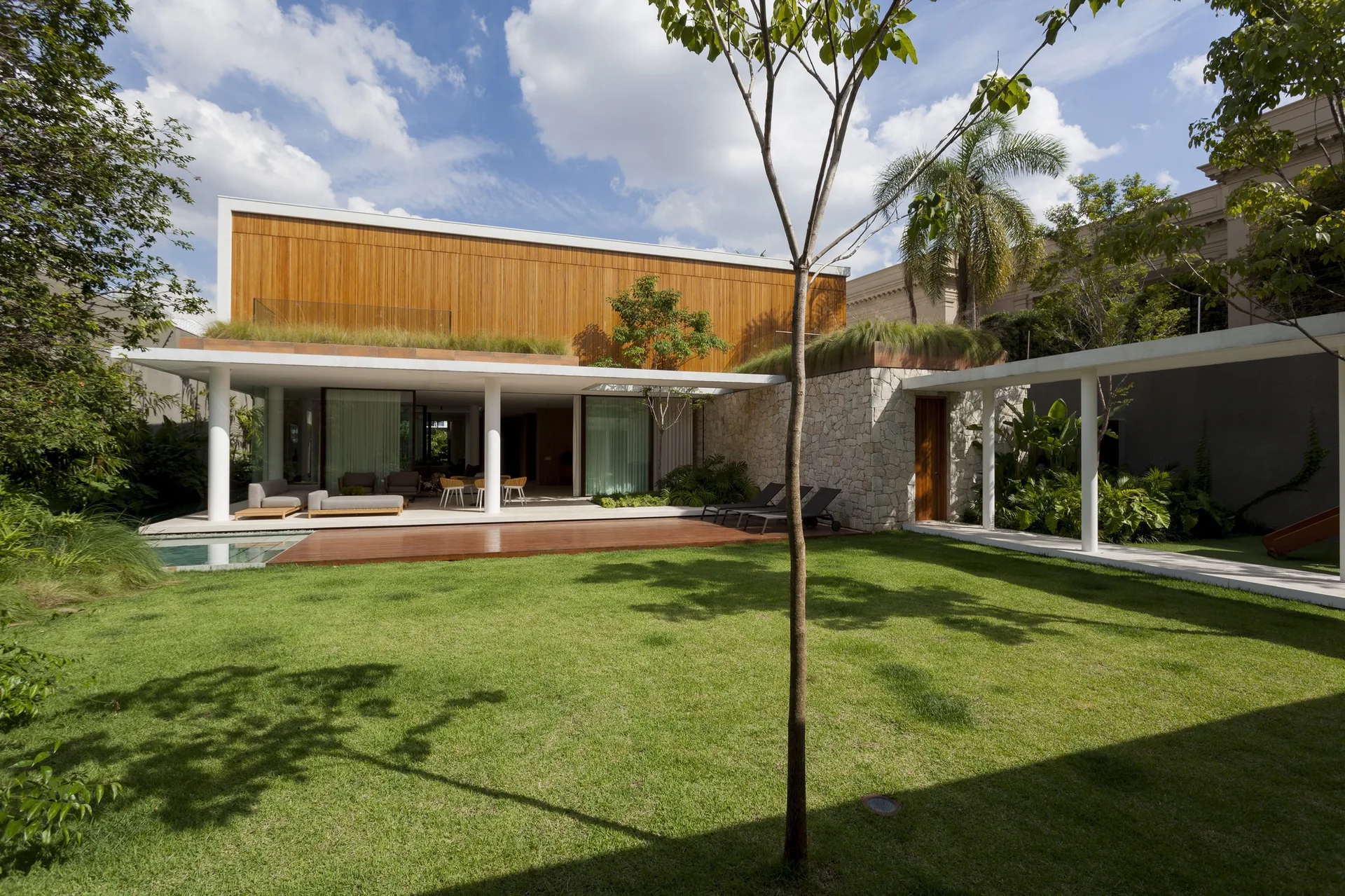 A modern residential backyard features a covered patio with white pillars, a natural stone wall, wood cladding, and a large green lawn.