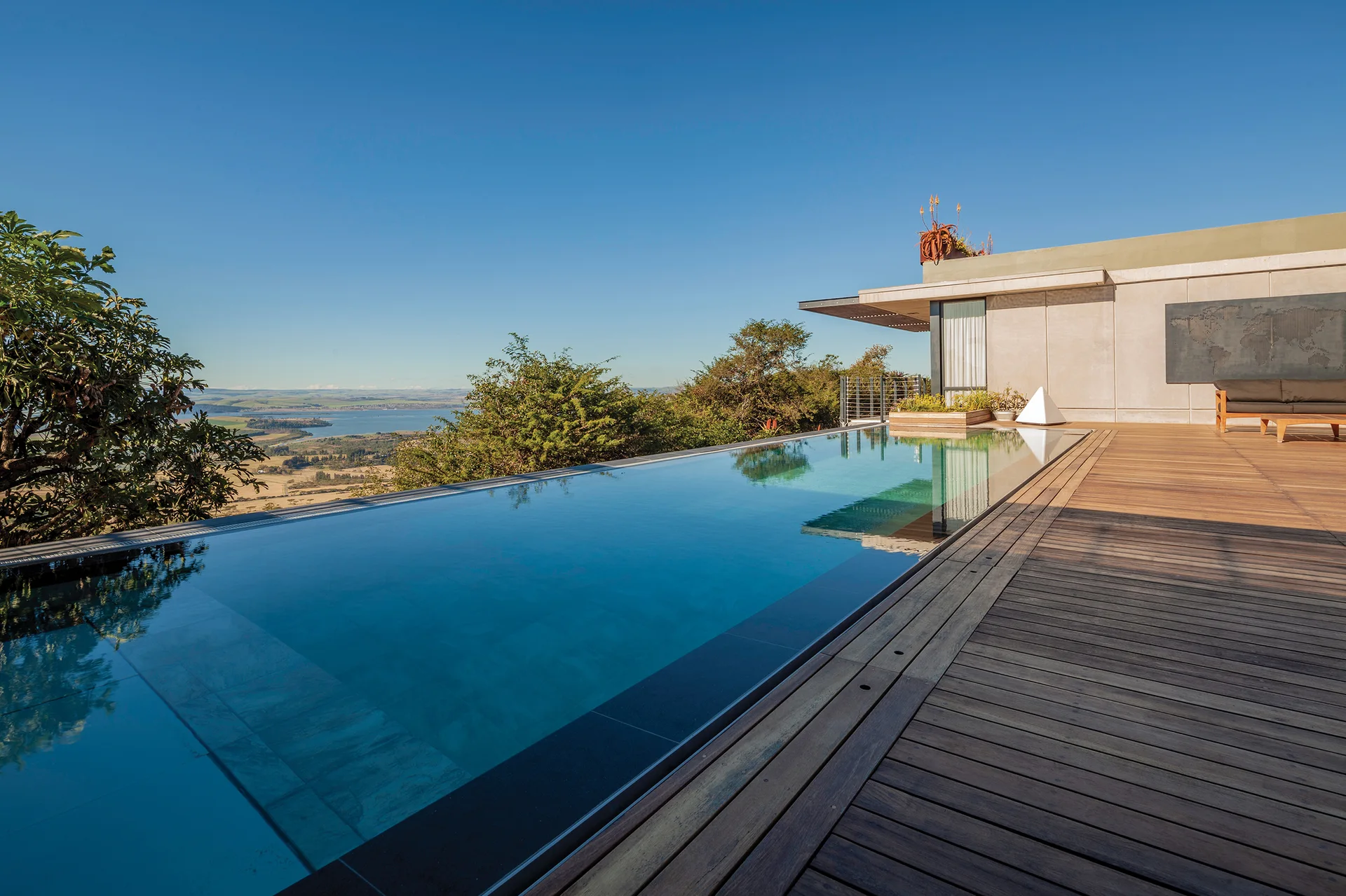 An infinity edge pool sits flush with a wooden plank deck, overlooking a green valley under a clear blue sky.