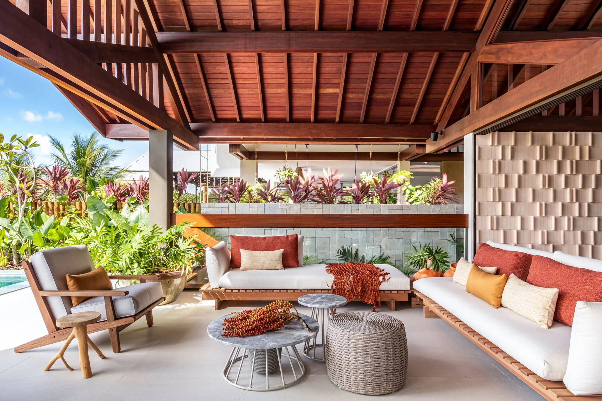 A covered patio lounge with wooden sofas, gray and orange cushions, nesting stone tables, and a high exposed timber ceiling.