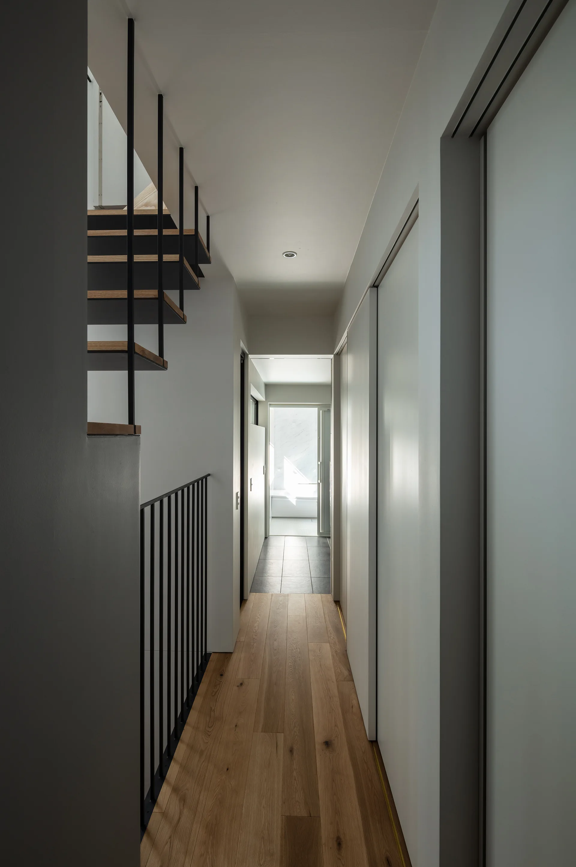 A minimalist white hallway features light wood plank flooring and an open-riser staircase with thin vertical black metal railings.