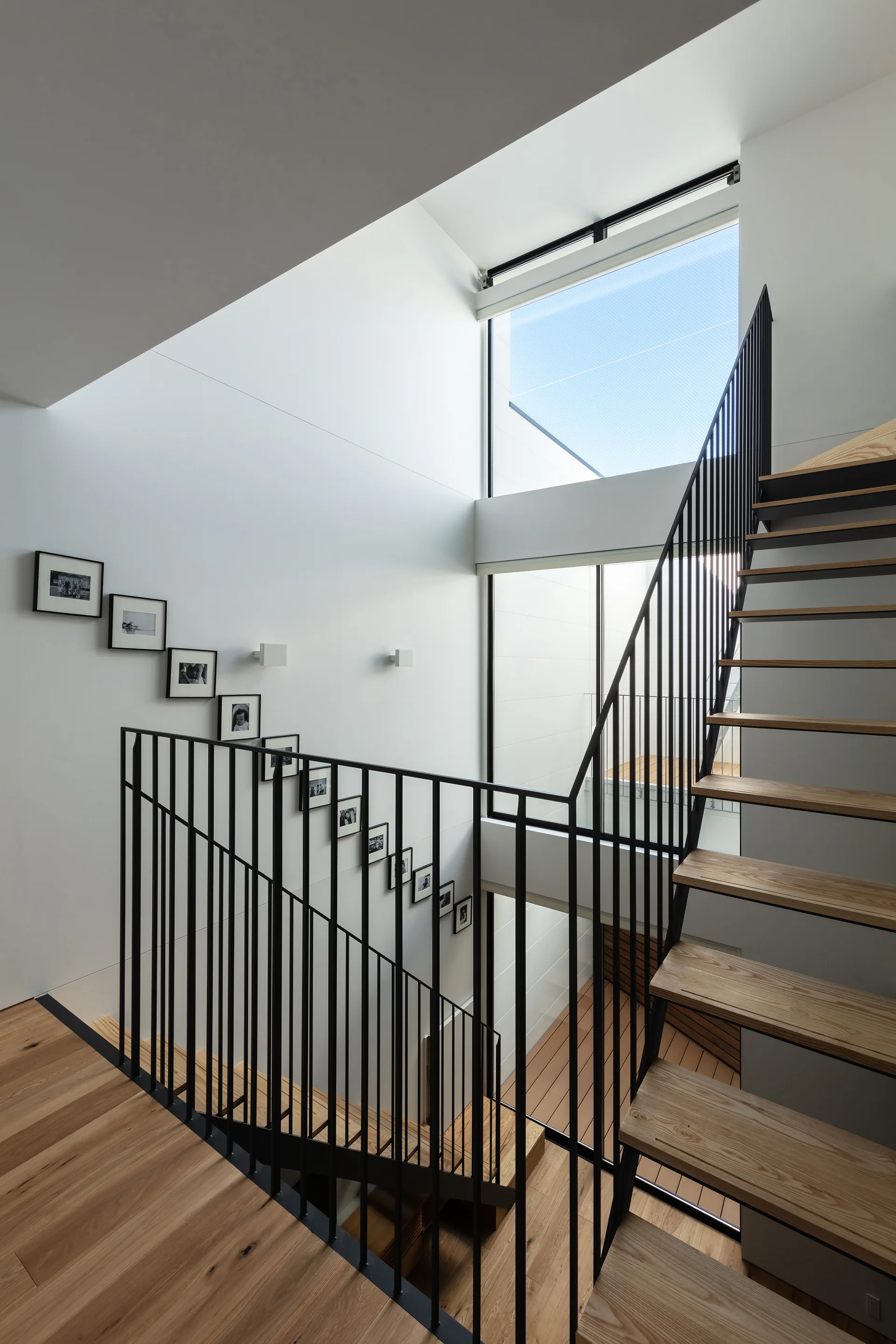 A modern staircase with light wood treads and thin black metal railings is set against white walls featuring a diagonal row of small framed photos.