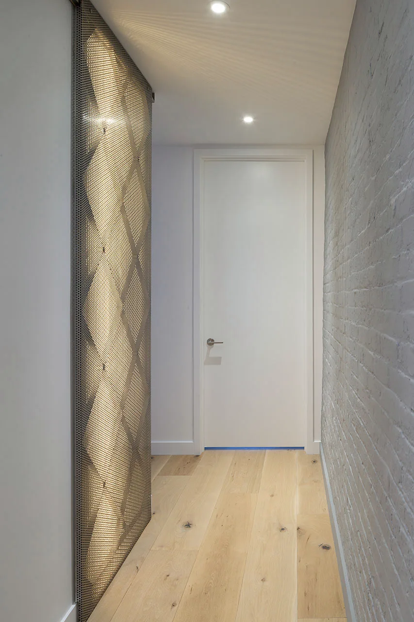 A narrow hallway features light wood floors, a painted white brick wall, a white door, and a tall backlit perforated metal screen.