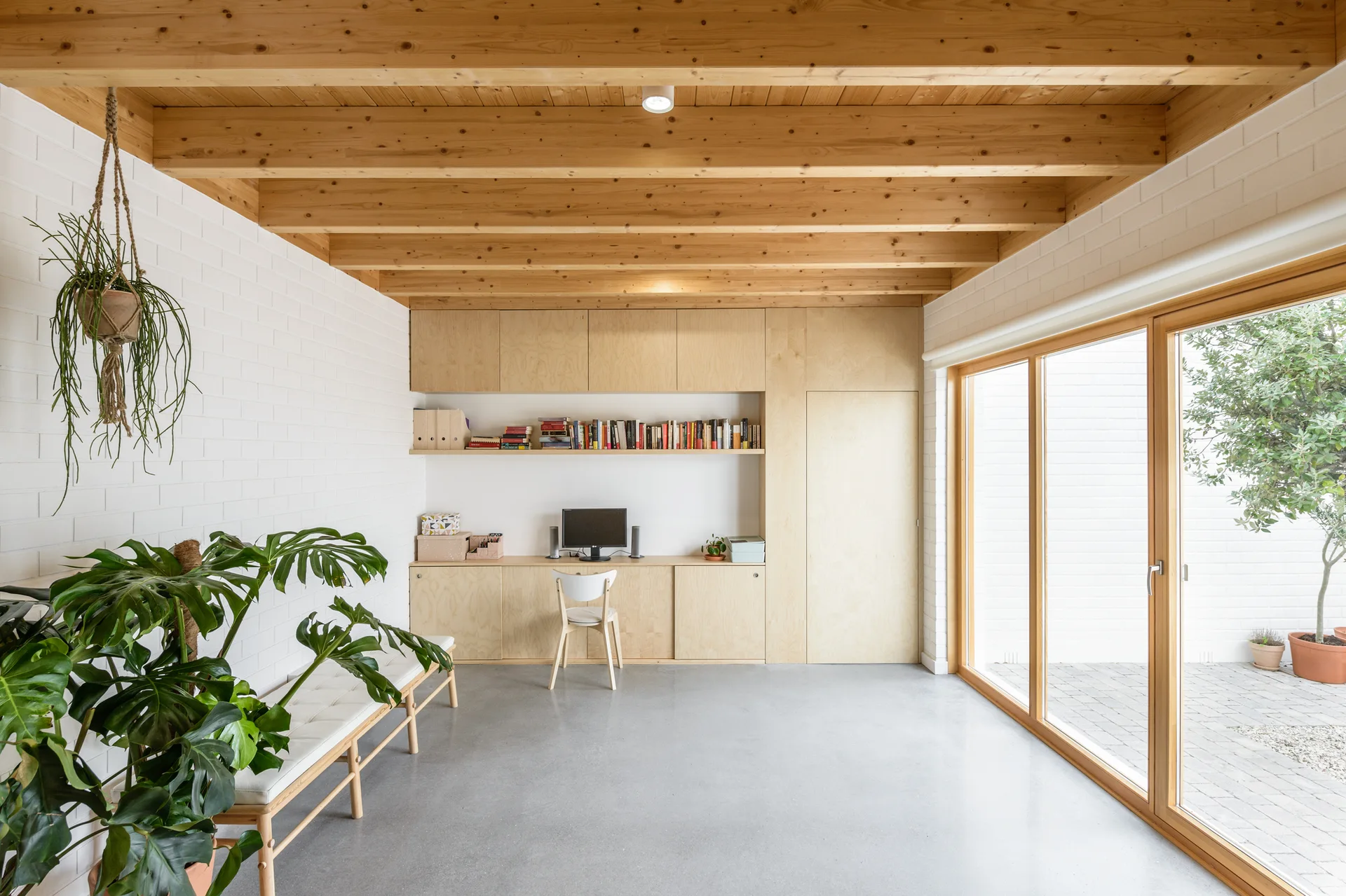 A minimalist home office featuring white painted brick walls, an exposed wood ceiling, a built-in wooden desk, and a large sliding glass door.