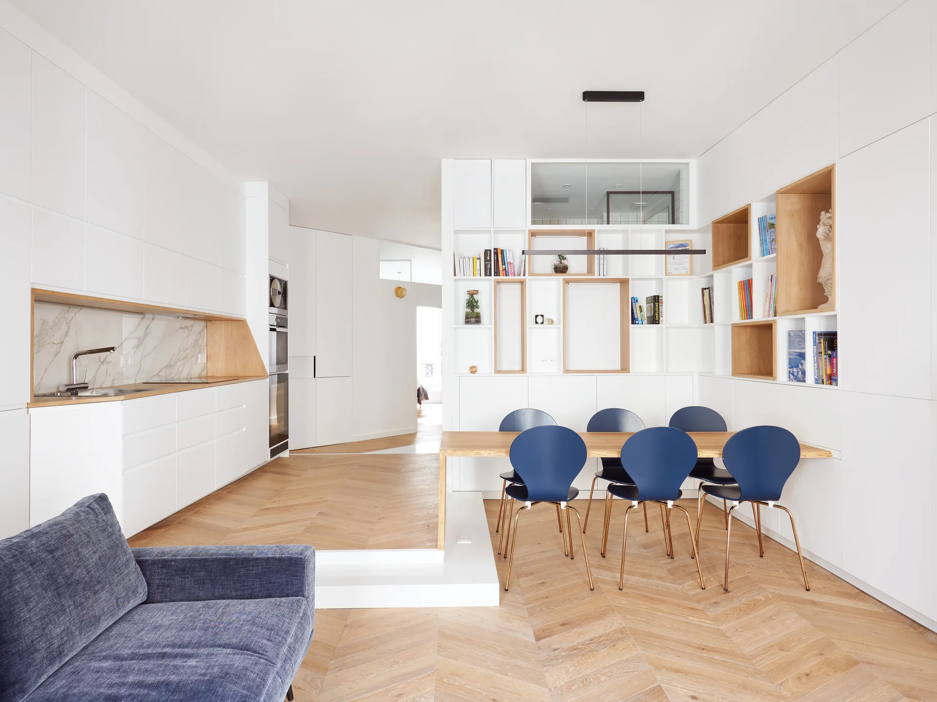An open-plan white kitchen and dining space with herringbone wood floors, a built-in shelving wall, and blue chairs around a light wood table.