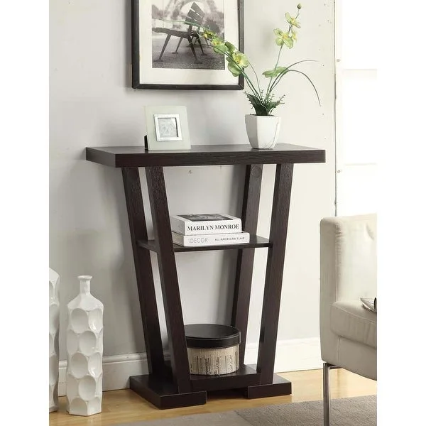 A dark brown wood console table with angled geometric legs holds a white orchid vase, standing against a grey wall with framed art.