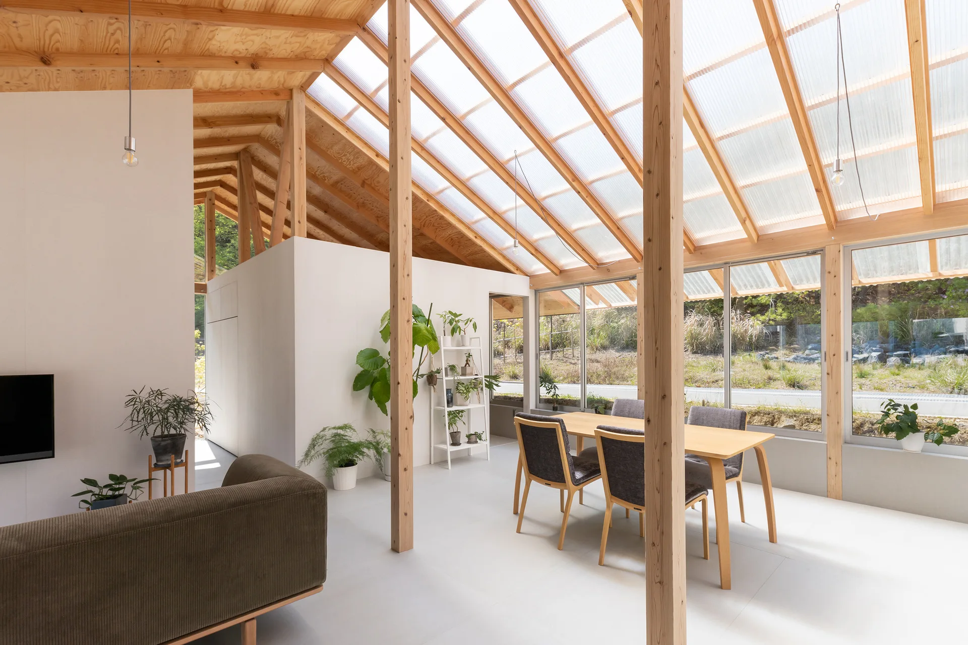An open-plan room features a wooden dining table with grey chairs under a sloped translucent roof with exposed timber framing.
