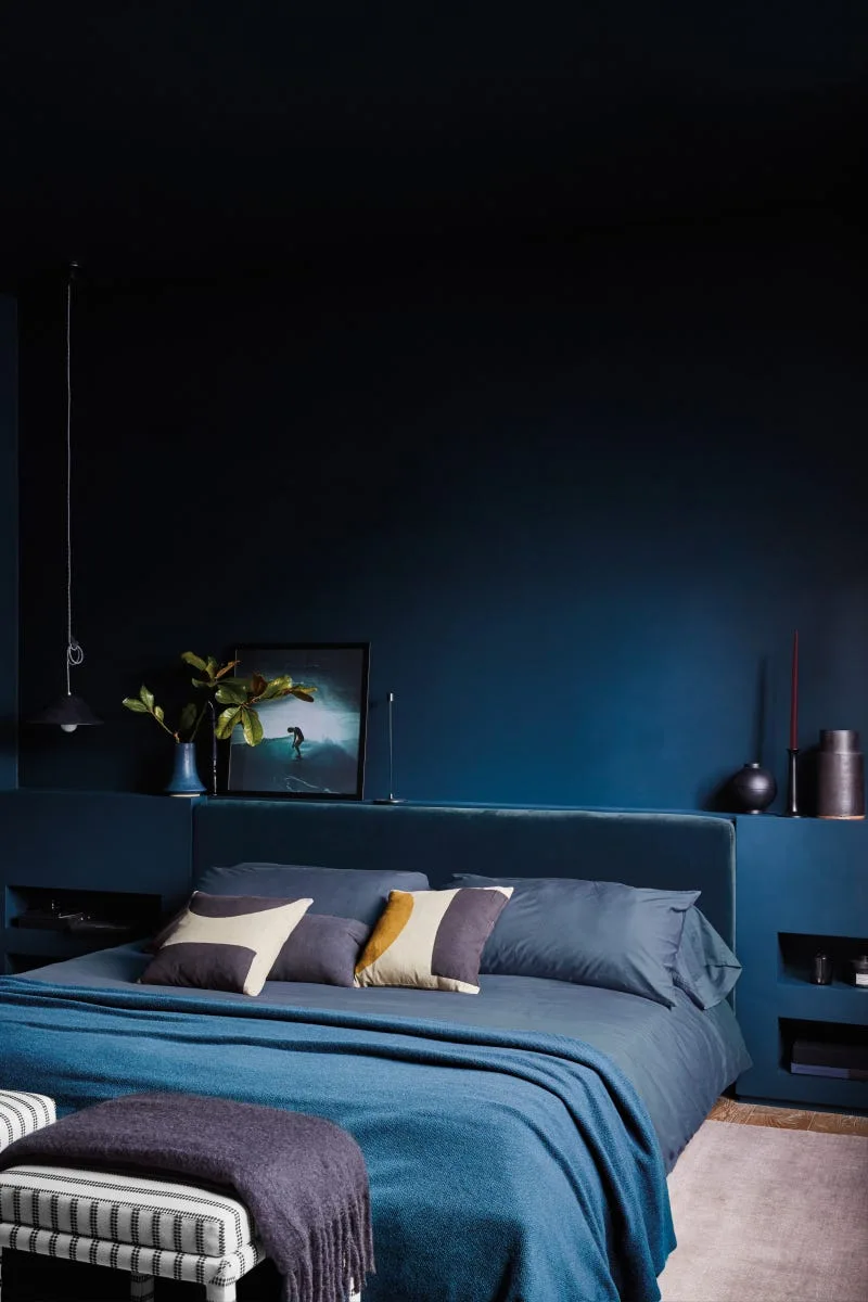 A dark blue bedroom featuring a built-in headboard storage ledge, matching navy bedding, and a striped bench at the foot of the bed.