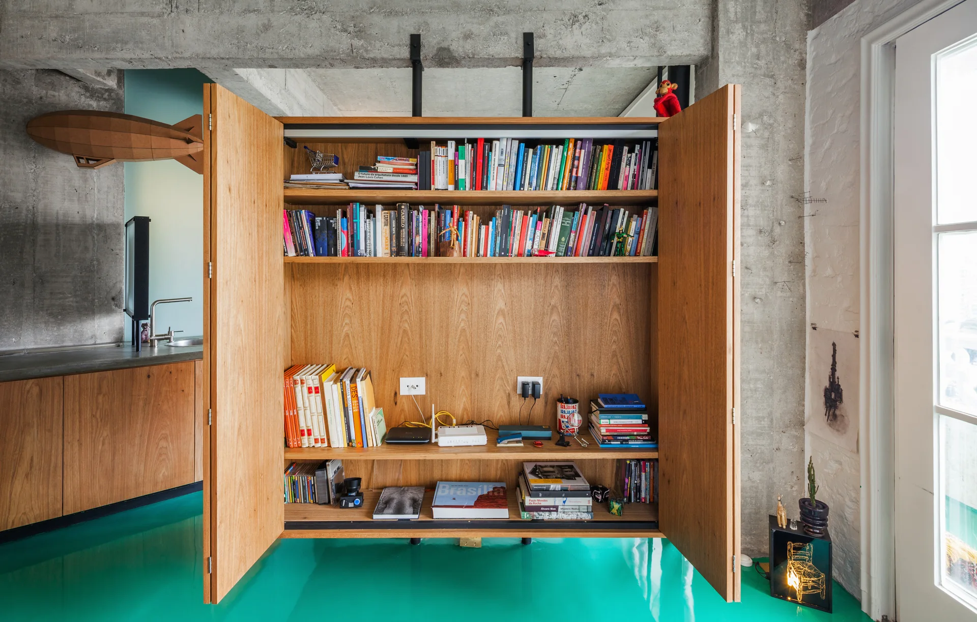An open wood bookcase with books and electronics sits on a glossy turquoise floor against raw concrete walls.