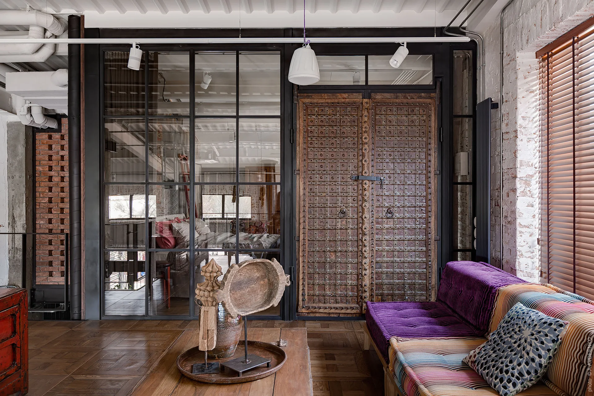 An eclectic loft space featuring a black steel-framed glass partition wall, studded antique wood doors, and a purple tufted sofa.