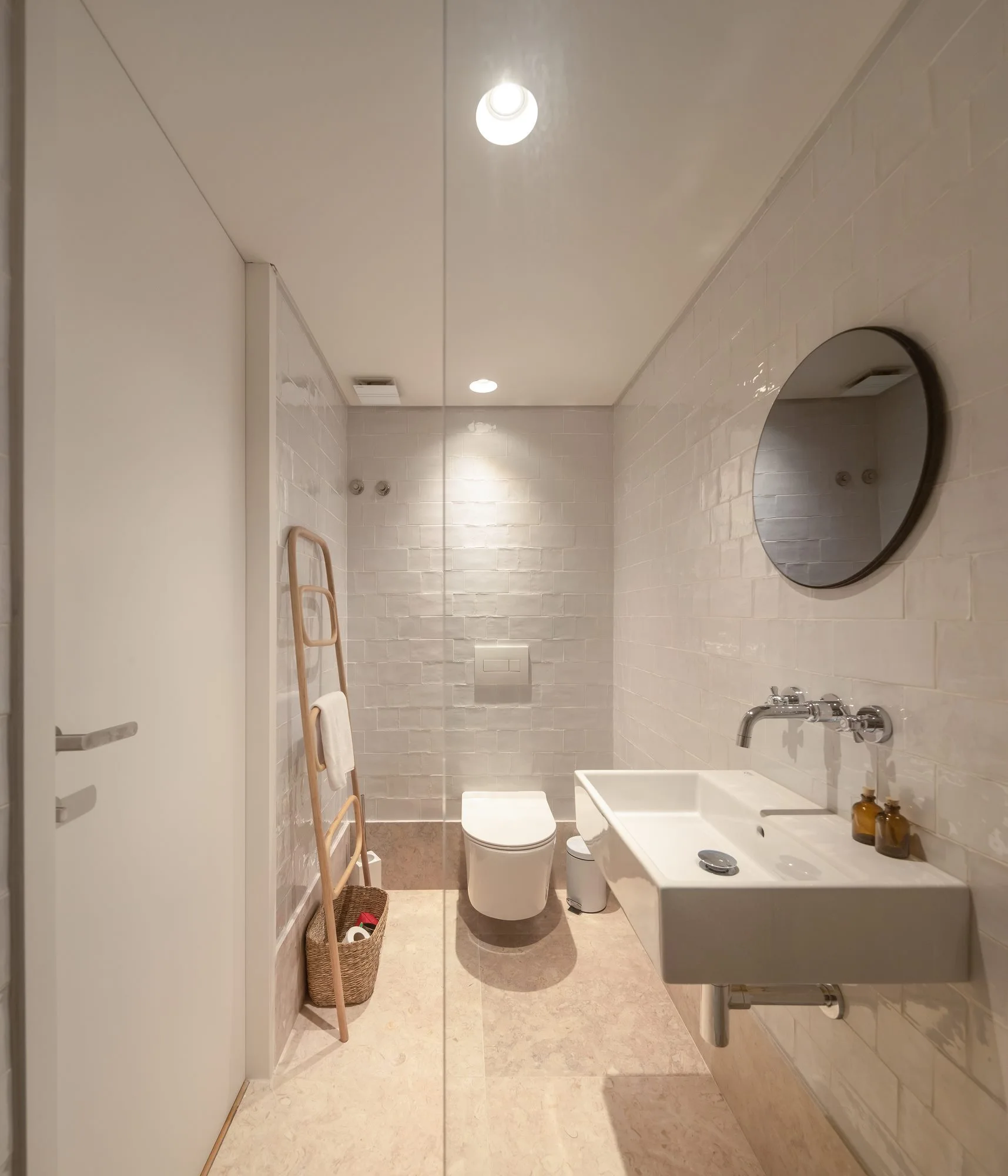 A narrow modern bathroom featuring glossy white subway tile walls, a wall-mounted sink, a wall-hung toilet, and a wooden towel ladder.