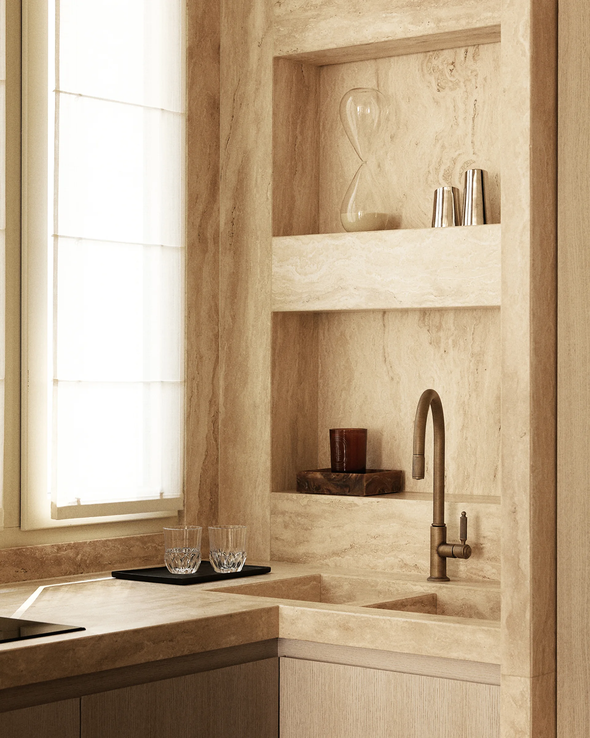 A kitchen corner features an integrated beige stone sink with a brass faucet, built-in recessed stone shelves, and a window with a white Roman shade.